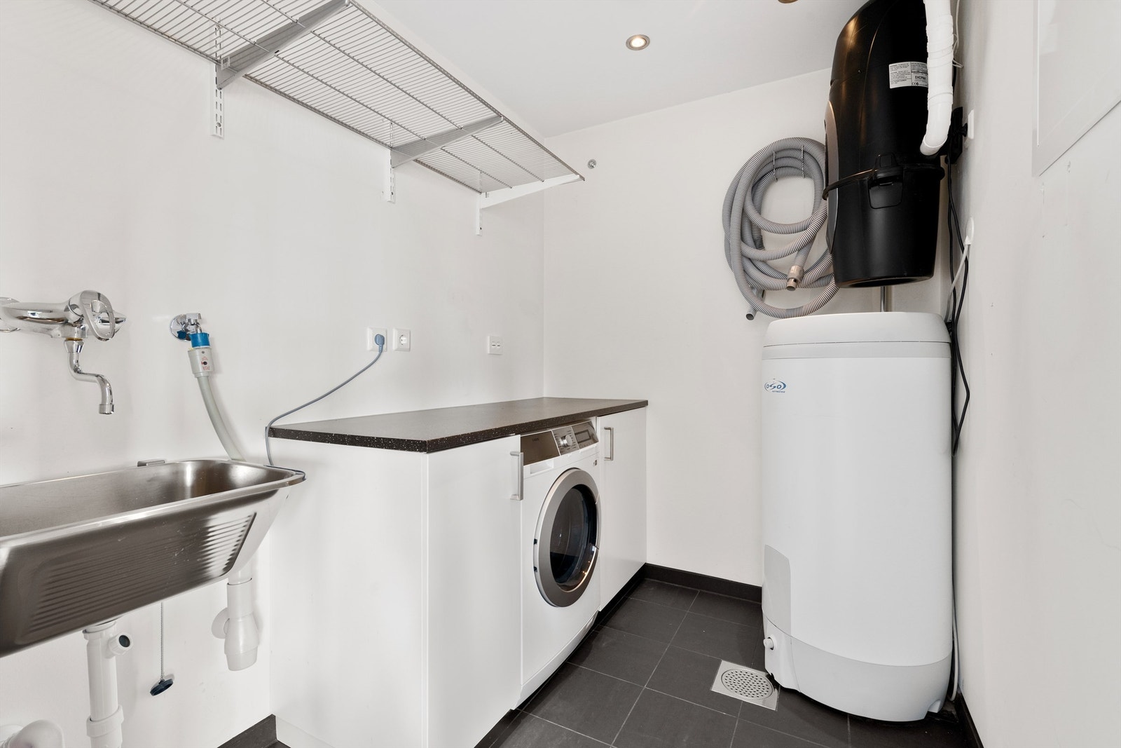 Praktisk vaskerom med flislagt gulv, utslagsvask og opplegg for vaskemaskin. Her er også sentralstøvsuger og varmtvannsbereder plassert. Galleribilde