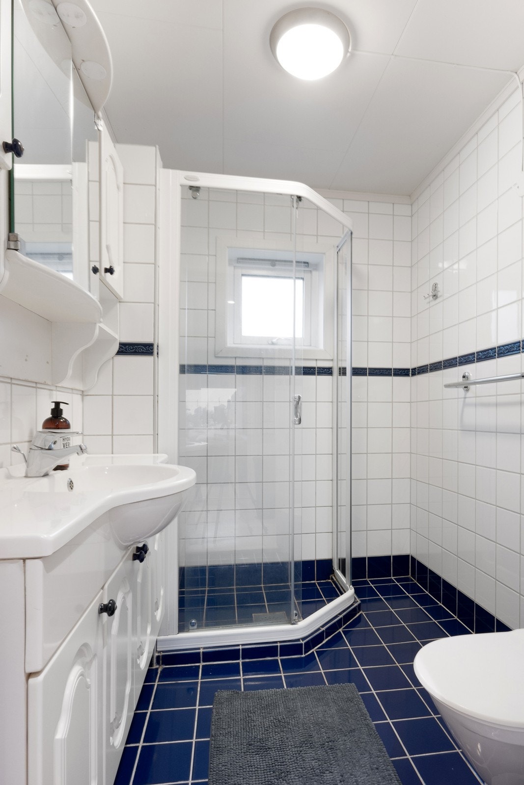 Badet er utstyrt med dusjhjørne og gulvmontert toalett, samt en helstøpt servant med oppbevaringsplass både i underskap og i speilskapet over vasken. Galleribilde