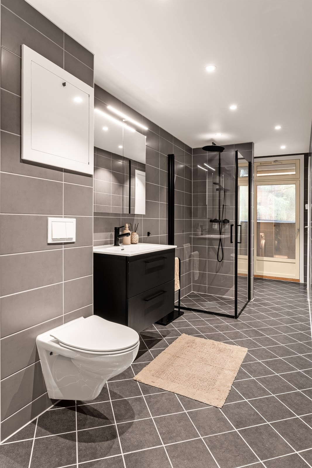 Innredning og garnityr på bad fremstår med veggmonterte dusjdører, servant med underskap, høyskap, speilskap med lys over samt veggmontert wc. Her er det også opplegg for vaskemaskin. Galleribilde