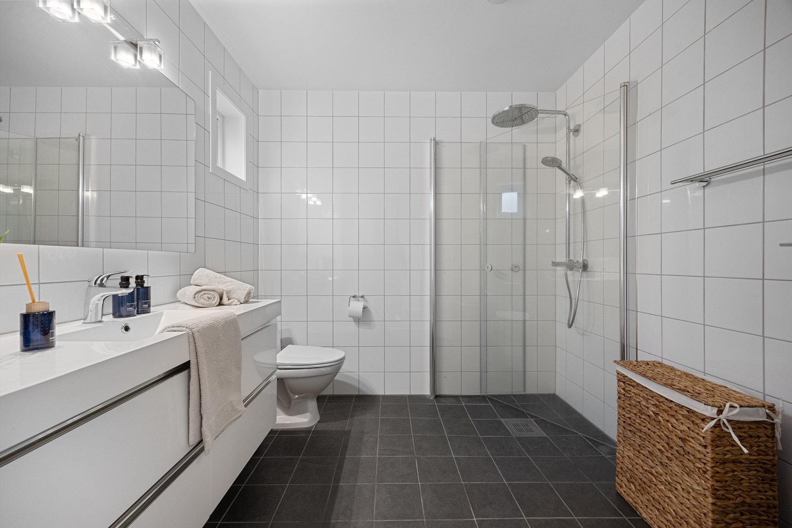 Badet er romslig og har varmekabler i gulv, heldekkende servant med skuffer, speil med lys, veggskap, gulvmontert wc og praktisk hjørnedusj med svingbare glassdører Galleribilde
