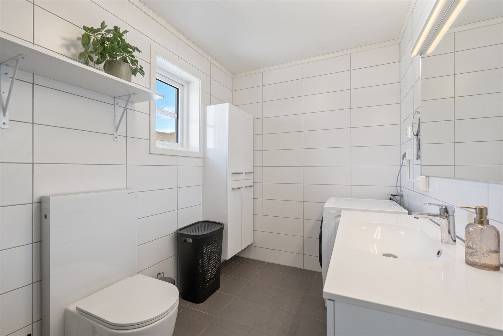 Badet er helfliset med mørke gulvfliser og hvite veggfliser. Det er opplegg for vaskemaskin. Galleribilde