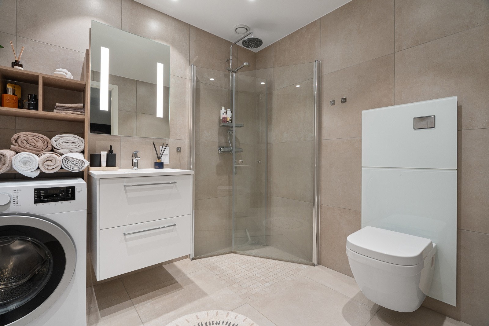 Lekkert bad med store beige fliser og utstyrt med praktisk vegghengt toalett, romslig dusjhjørne og plass for vaskemaskin. Galleribilde