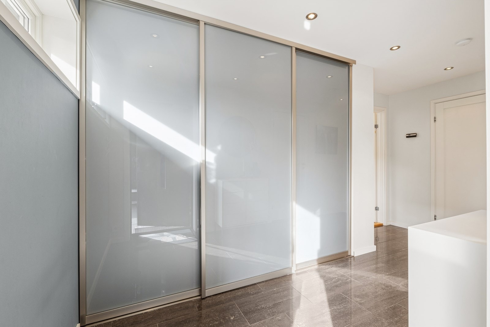 Entreen har fliser på gulv, lysmalte veggflater og spotter i taket. Stor skyvedørsgarderobe gir rikelig med oppbevaringsplass både for yttertøy og sko. Galleribilde