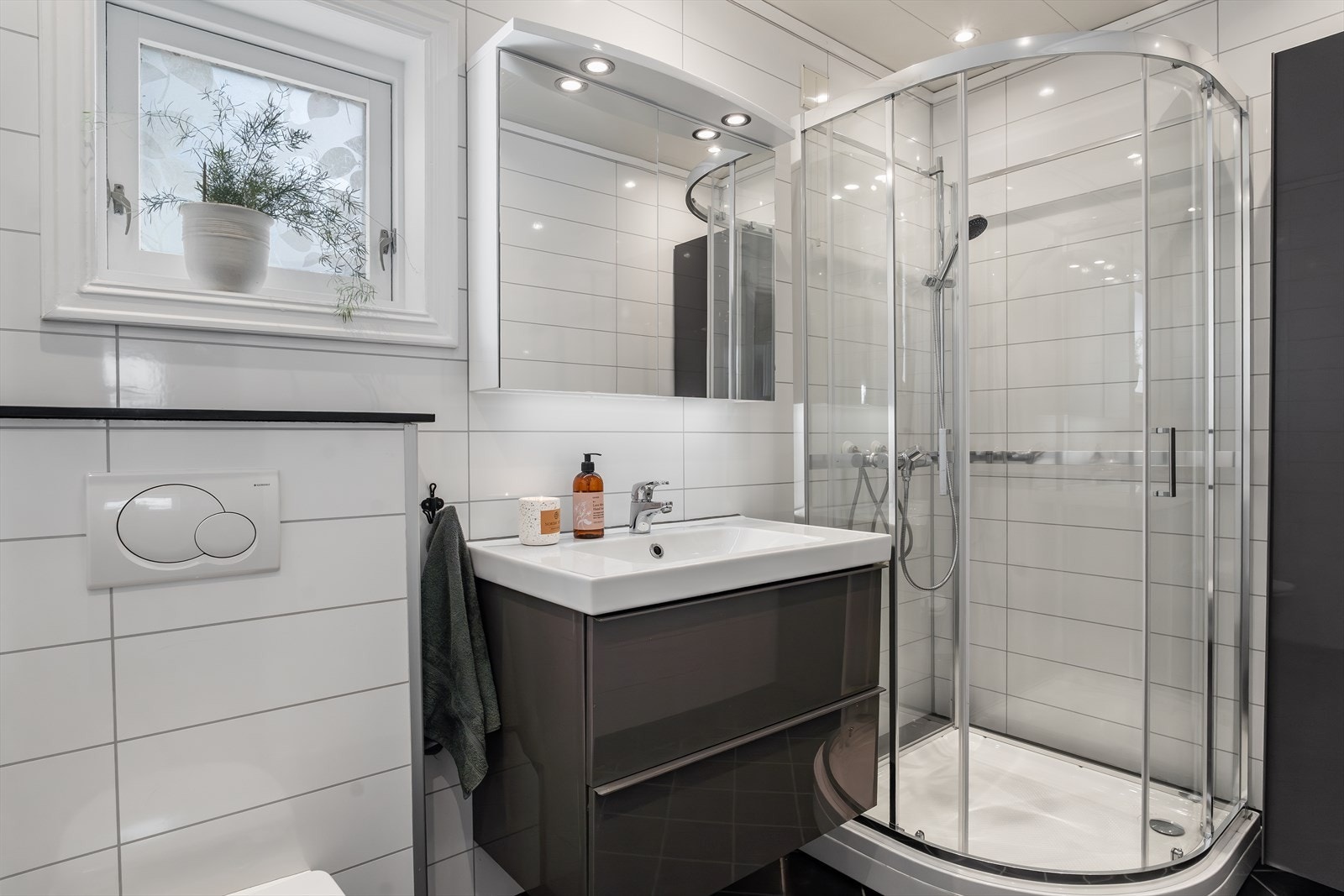 Badet har fliser på gulv og baderomsplater på veggene, og er utstyrt med vegghengt toalett, dusjkabinett, grå baderomsinnredning, servant med oppbevaring og belysning. Galleribilde