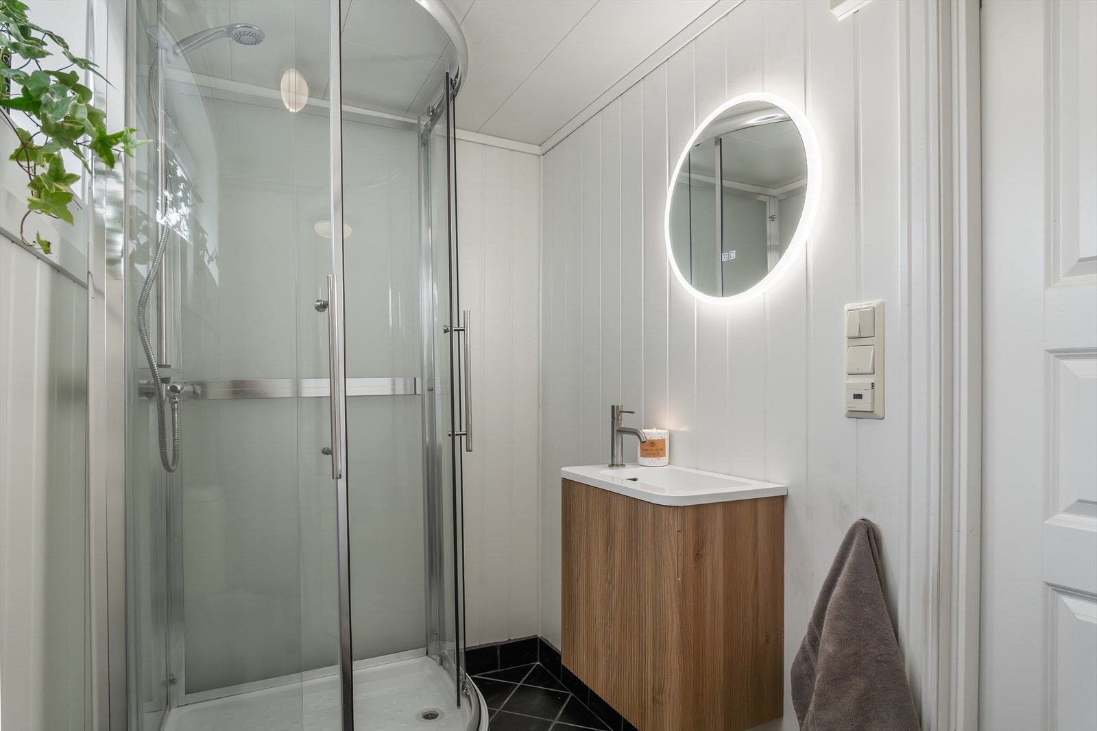 Bad med fliser på gulv og panel på vegger, utstyrt med dusjkabinett, toalett, servant og speil med belysning. Galleribilde