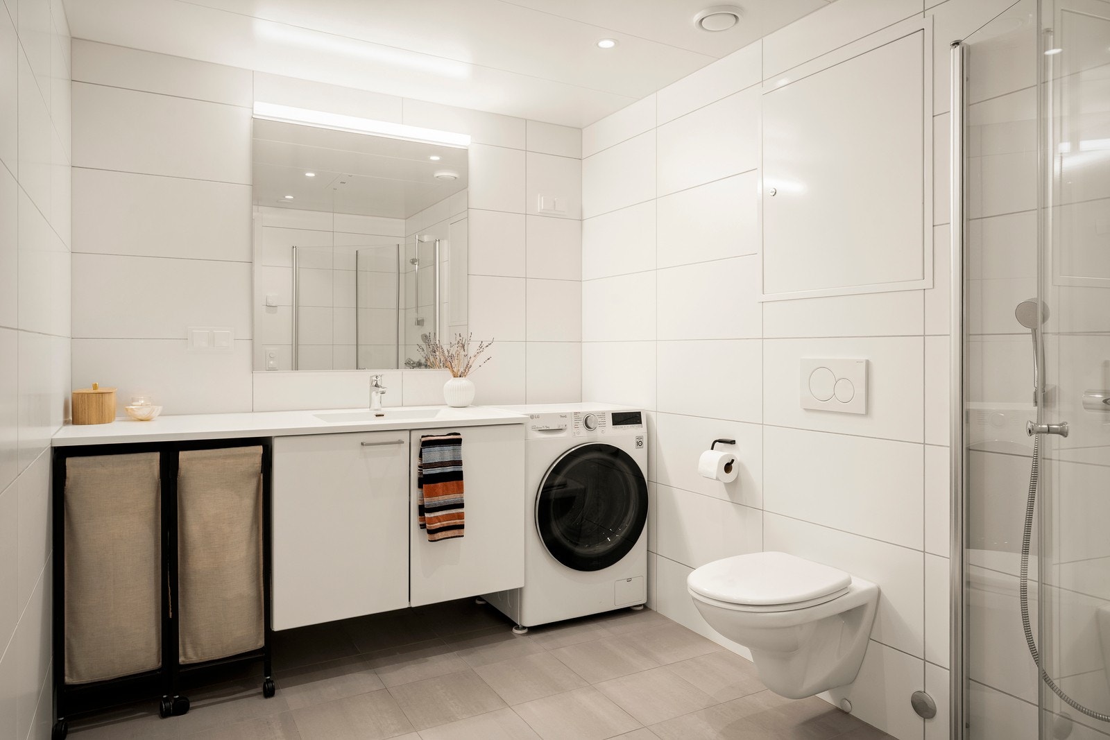 Baderomsinnredning bestående av heldekkende servant med slette lyse fronter, servant armatur og overhengende speil med belysning. Dusjen er plassert i hjørnet og har dører av glass. Videre er det montert vegghengt toalett og opplegg for vaskemaskin. Galleribilde