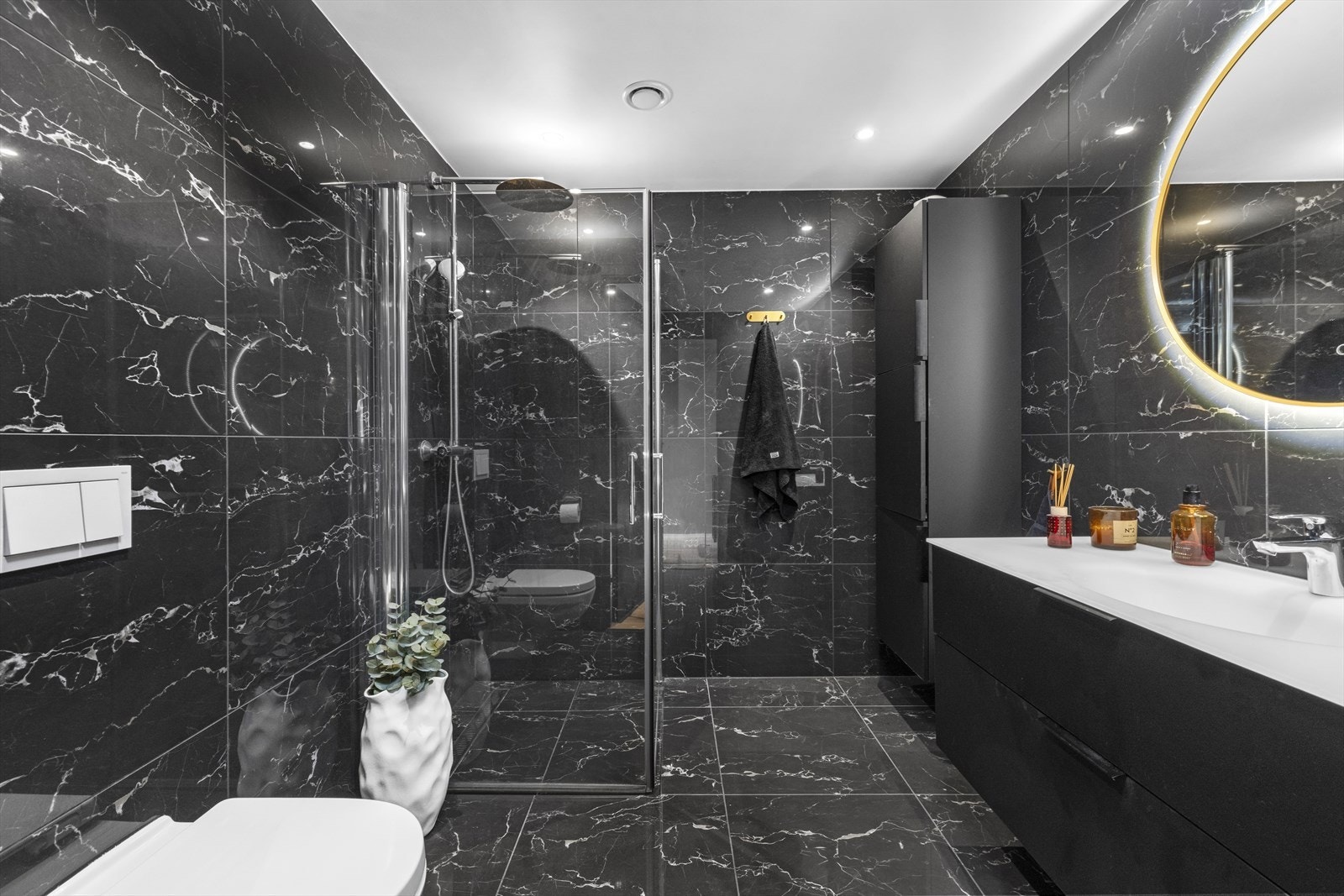 Badet er utstyrt med heldekkende servant over skuffeinnredning, overliggende speil med belysning, vegghengt skap, dusjhjørne med innfellbare glassvegger, samt vegghengt toalett. Galleribilde