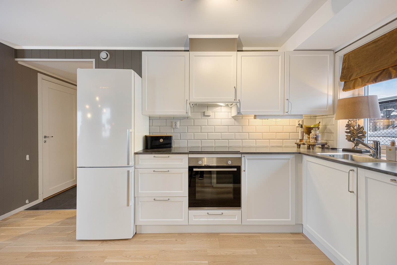 Kjøkkenet er oppført i en funksjonell L-form med god arbeidsflyt og skapplass. Innredningen har hvite, profilerte fronter, nedfelt oppvaskkum og laminert benkeplate i mørkegrått. Galleribilde