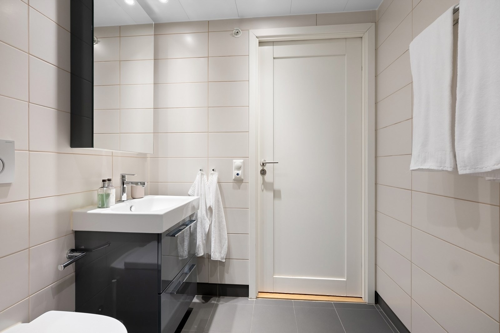 Badet inneholder veggmontert toalett, dusjhjørne med glassdører og to dusjhoder (håndholdt og regnfall) og baderomsinnredning med mørke, høyglanslaminerte skuffefronter, keramisk servant og ettgreps krummet armatur fra Grohe. Galleribilde