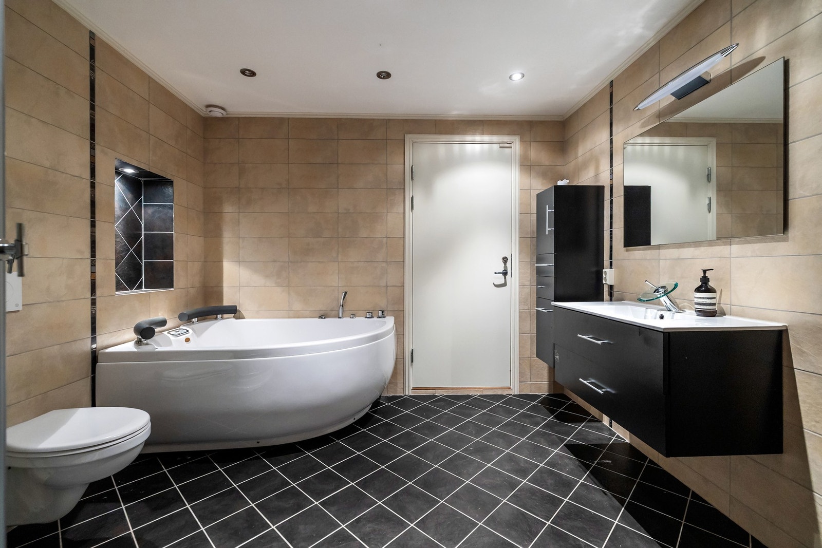 Badet i underetasjen har flislagte overflater med gulvvarme, og det er opplegg for vaskemaskin, som gjør hverdagen enkel og effektiv. Innredningen består av nedfelt servant i skuffeseksjon, spaeil, høyskap, badekar dusjkabinett og vegghengt toalett Galleribilde