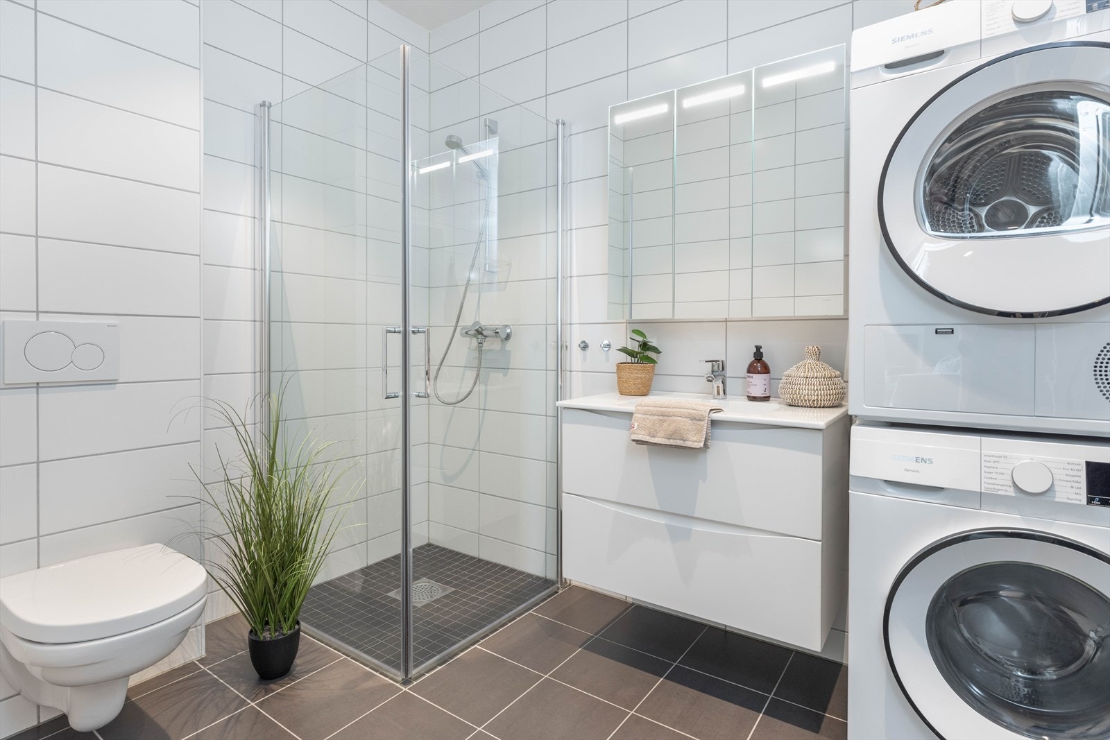 Pent og moderne helfliset bad, som er innredet med baderomsmøbel, speil med belysning, vegghengt toalett og dusjhjørne med innfellbare glassdører. Galleribilde