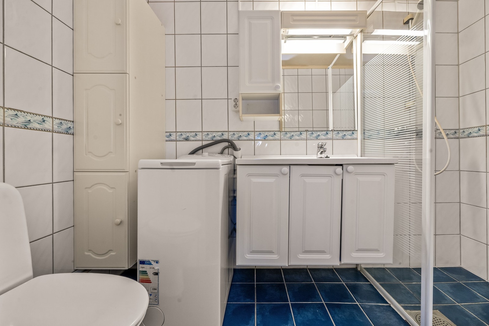 Leilighetens baderom er utstyrt med nedfelt servant, gulvfestet toalett, dusjhjørne og en vaskemaskin som medfølger salget Galleribilde