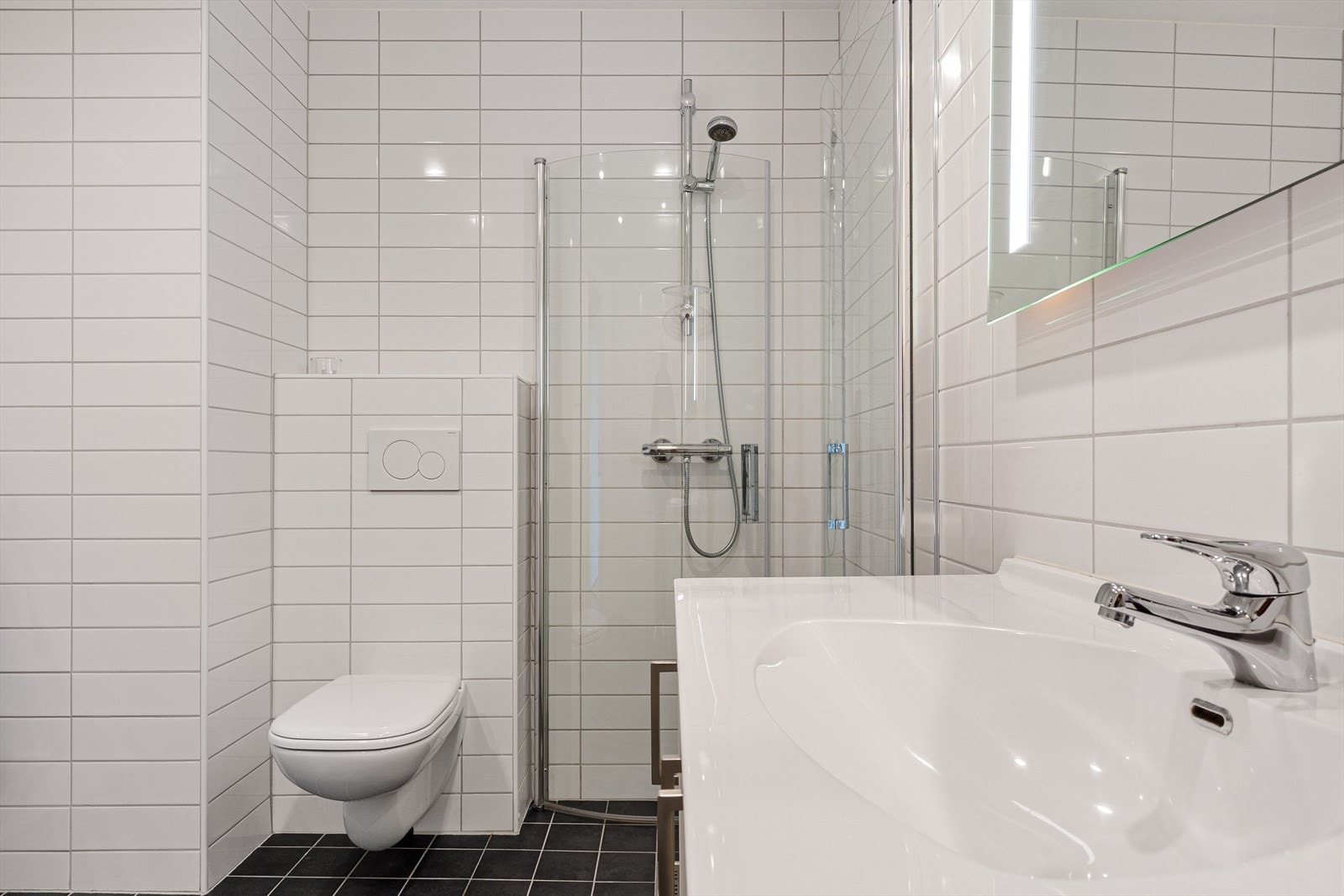 Badet er innredet med vegghengt toalett, dusj med innfellbare dusjdører og servantmøbel med speil og integrert belysning. Galleribilde