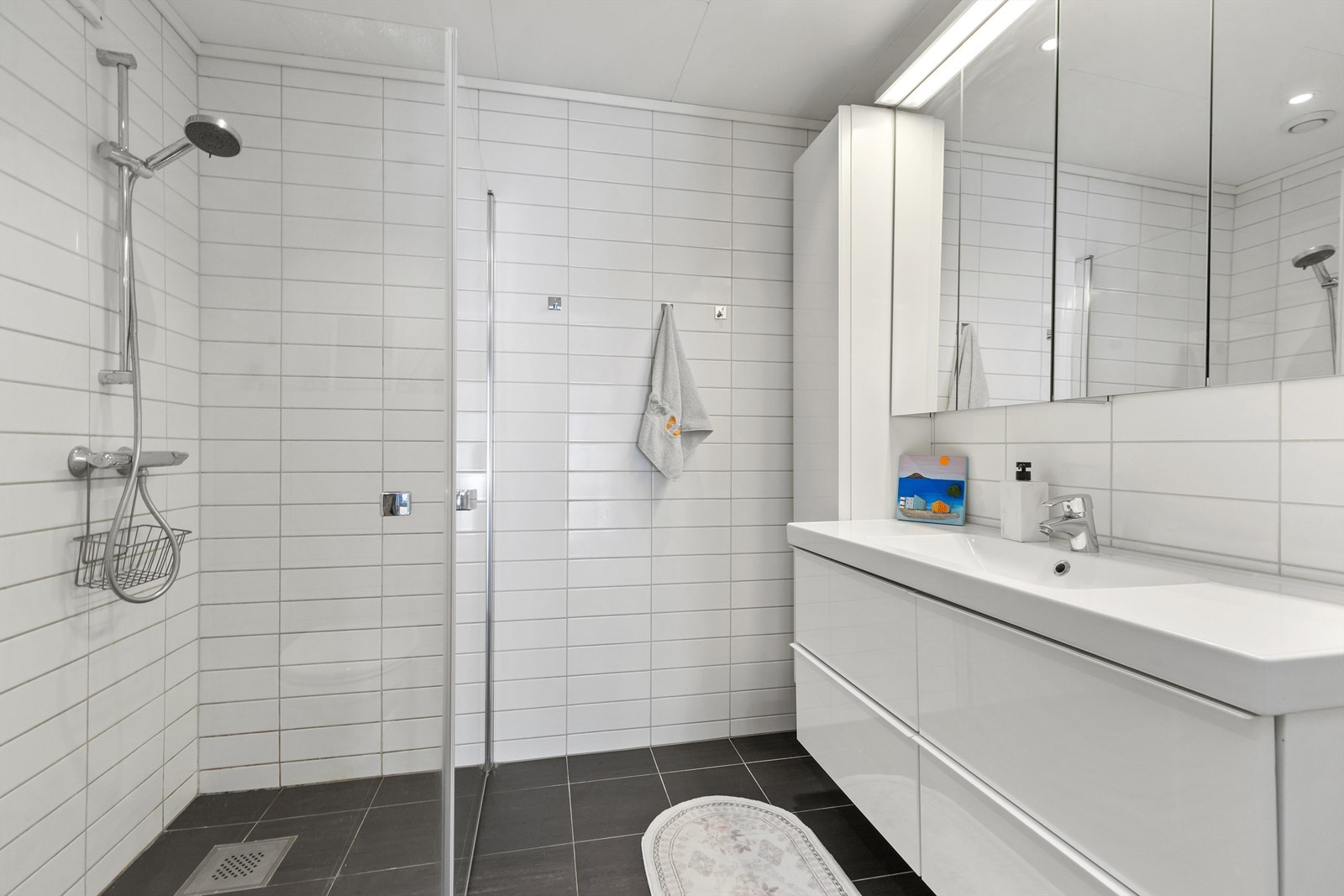 Badet er utstyrt med dusjhjørne med innfellbare dører, vegghengt toalett og servantinnredning. Galleribilde