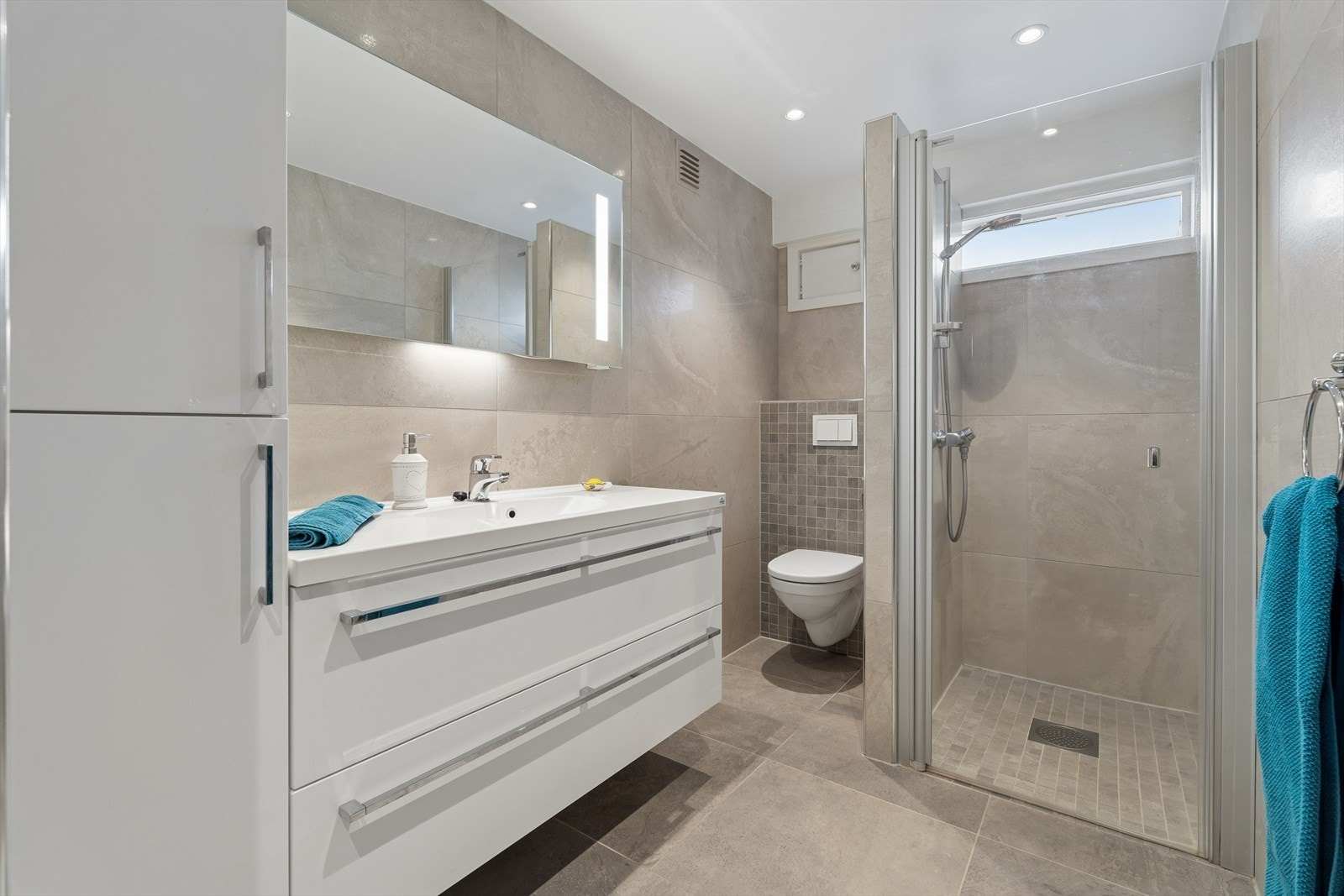 Dette badet er også fra 2015 med elektriske varmekabler - Rommet er innredet med servant i nedfelt innredning, veggmontert toalett og et romslig dusjhjørne med glassvegger. Galleribilde