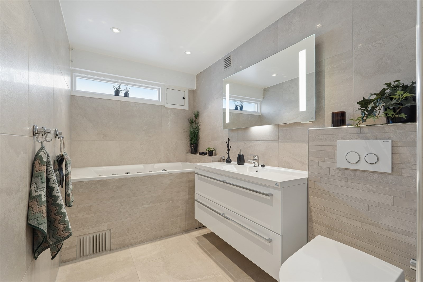 Rommet er innredet med en stilren baderomsinnredning med nedfelt servant, veggmontert toalett og et innbydende badekar. Galleribilde