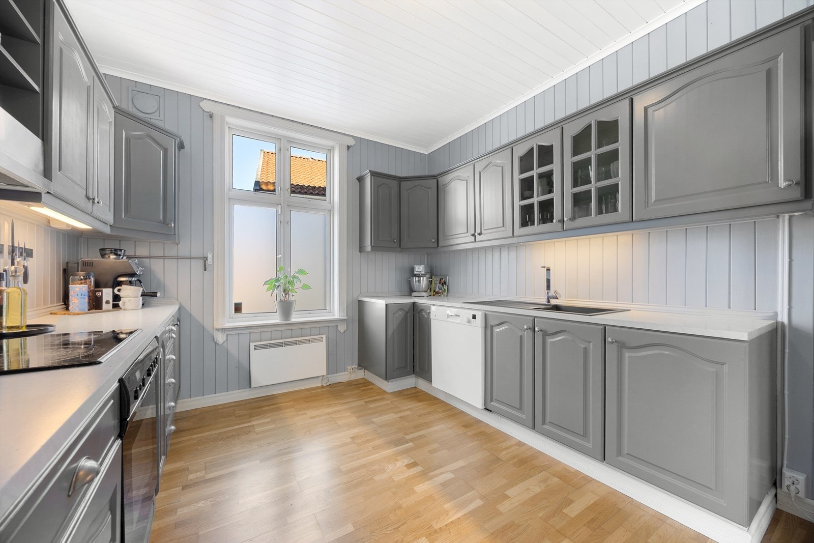 Kjøkkenet har parkettgulv, fint lysinnslipp og panelvegger malt i en delikat gråtone. Innredningen er oppført langs to vegger, med grå, profilerte fronter, vitrineskap og oppvaskkum nedfelt i en laminert benkeplate. Galleribilde