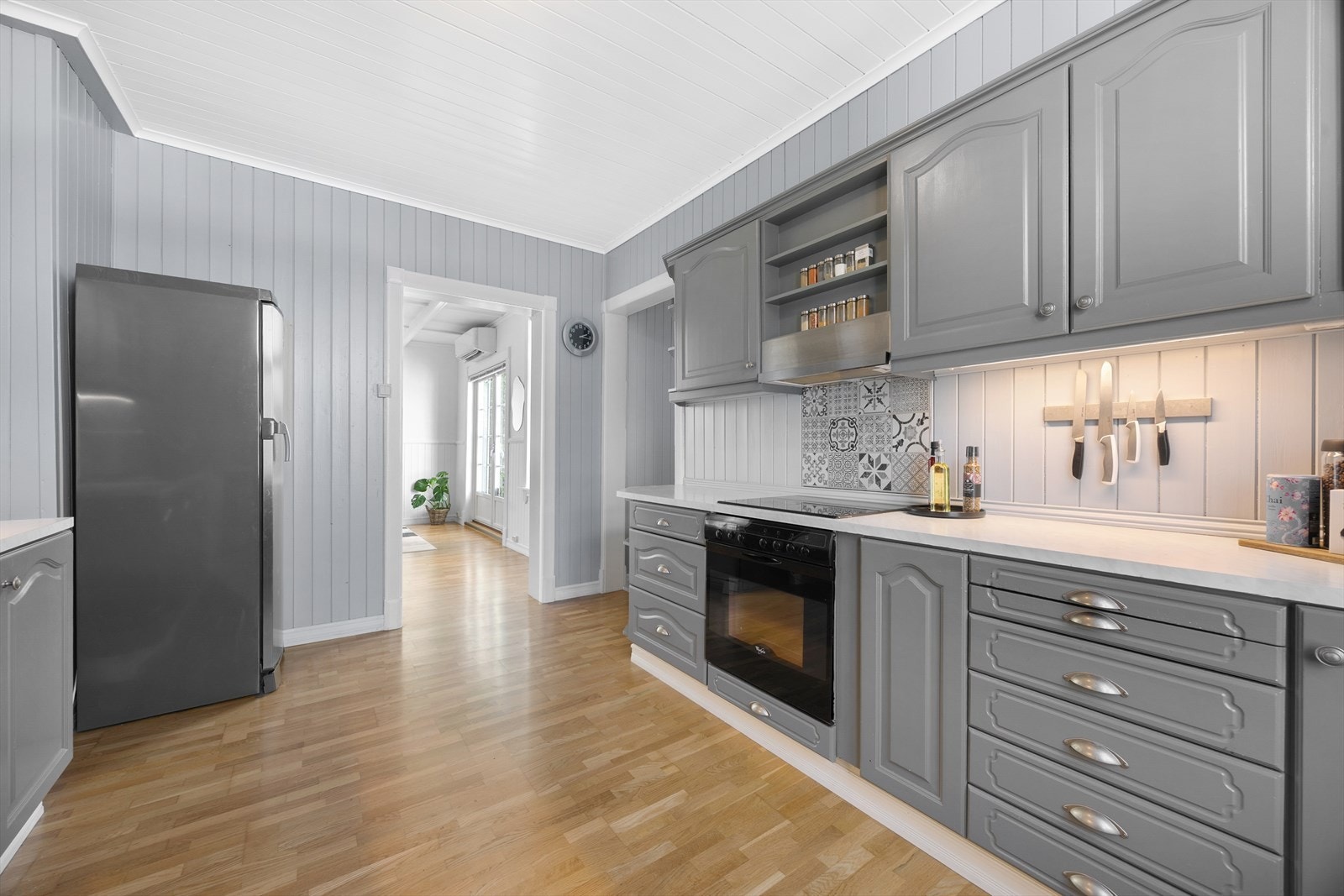 Belysning er montert over benken, og over kokesonen har kjøkkenet en ventilator, i tillegg til samme type fliser som entreen. Opplegg for oppvaskmaskin er etablert, og integrert i innredningen er det platetopp og stekeovn. Galleribilde