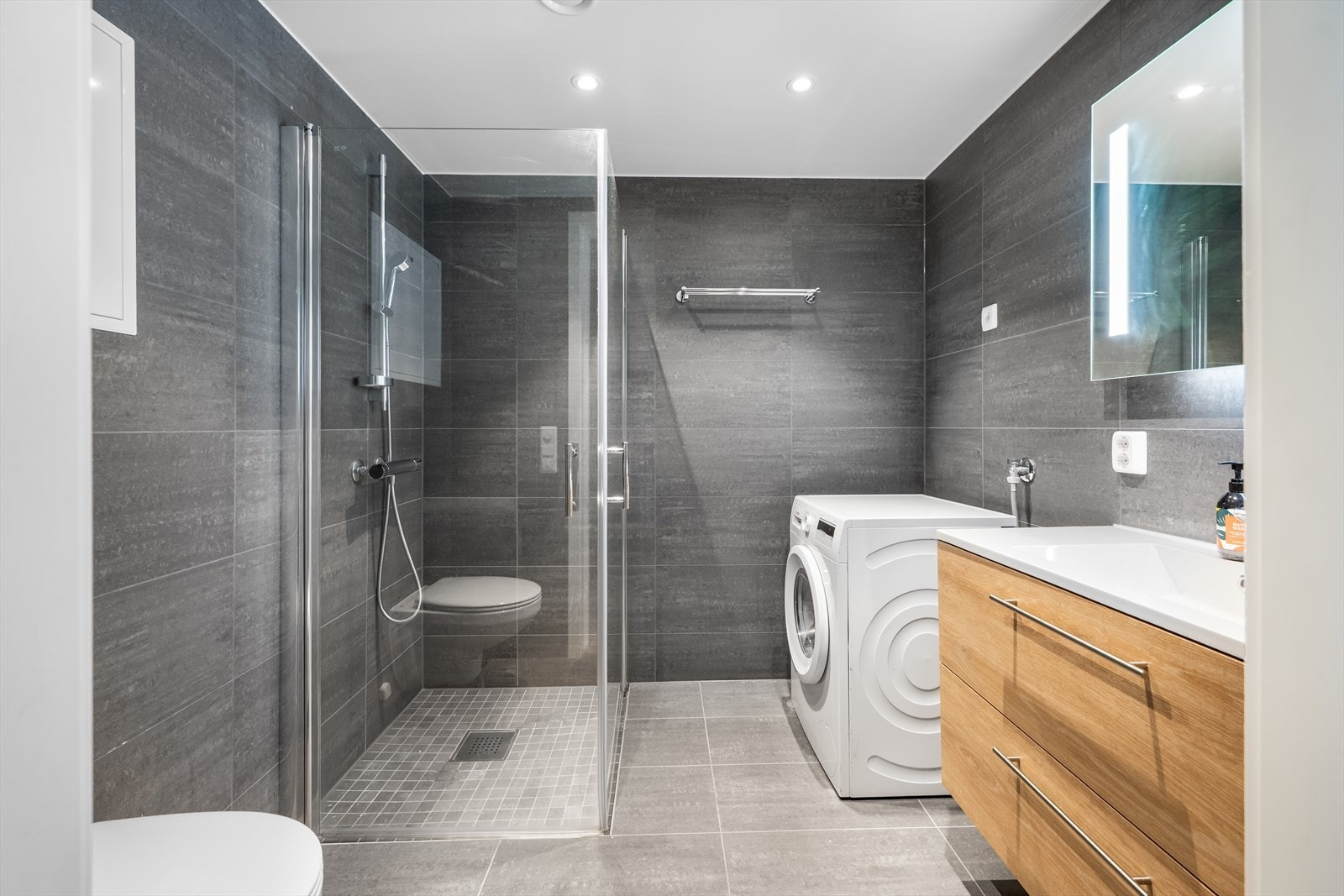 Badet er lekkert innredet med mørke veggfliser og lysere gulvfliser i en moderne gråtone Galleribilde