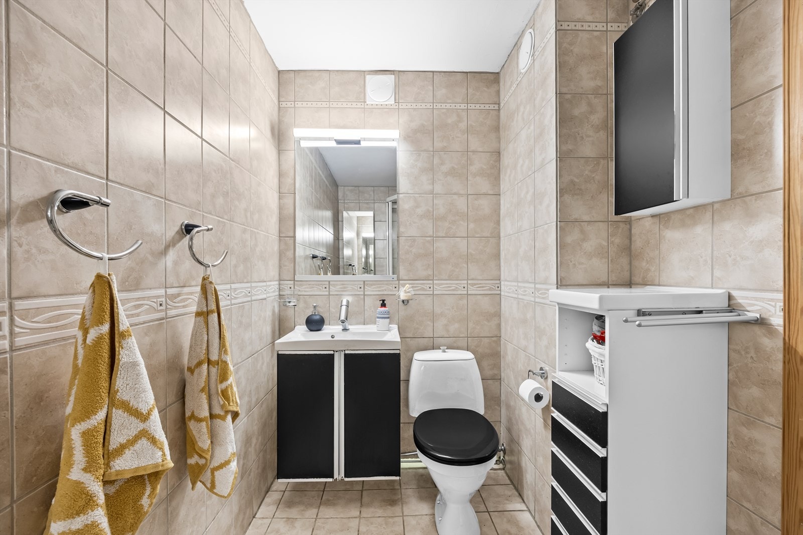 Romslig bad med inngang fra grovkjøkken. Badet har fliser på gulv og vegger og inneholder servant med underskap, toalett og dusjkabinett. Galleribilde