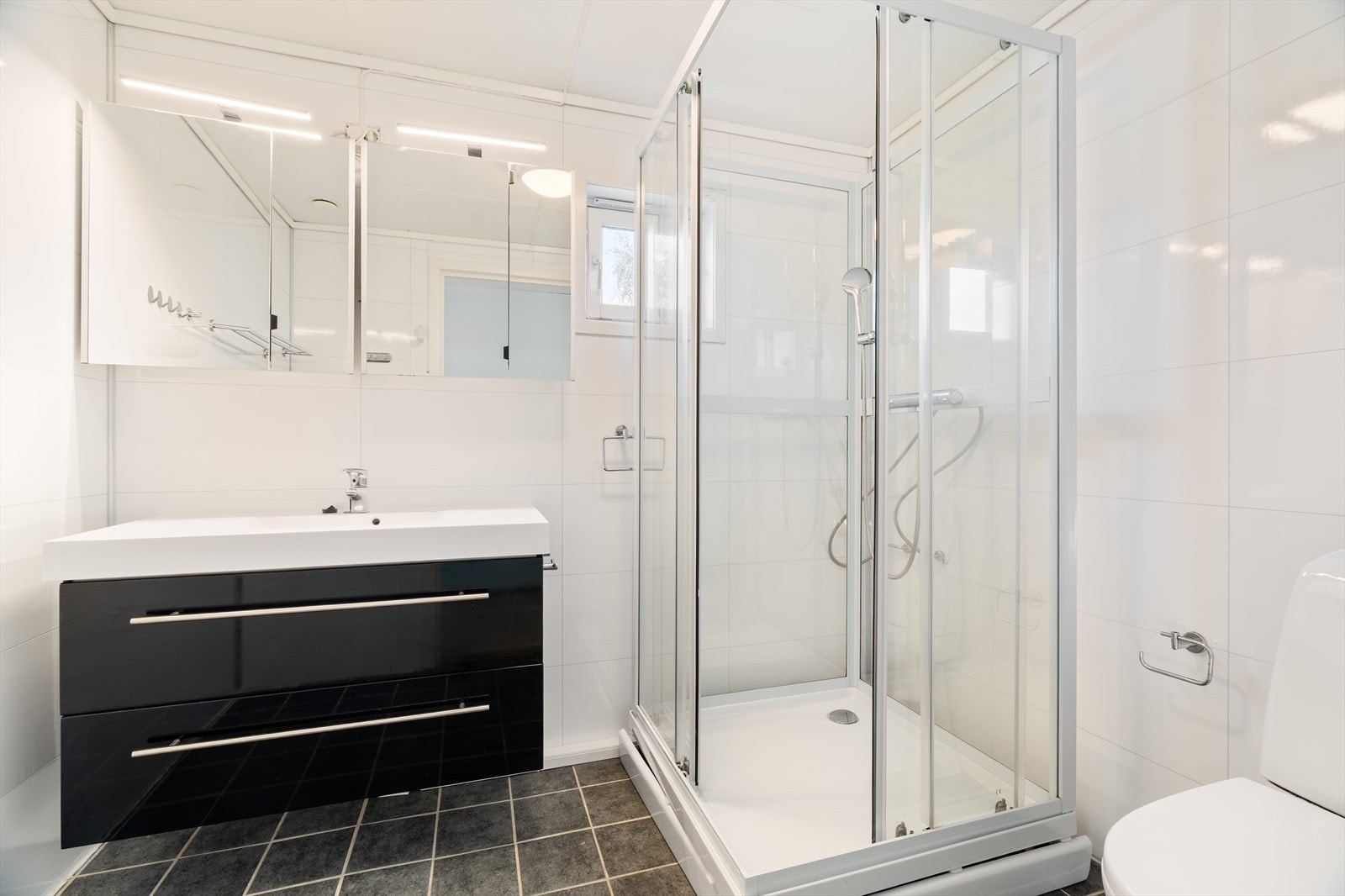 Badet er flislagt med hvite flis på vegger og mørk grå flis på gulvet. Det er innredet med servant og svart underskap, speil med oppbevaringsmuligheter, dusjkabinett og toalett. Galleribilde