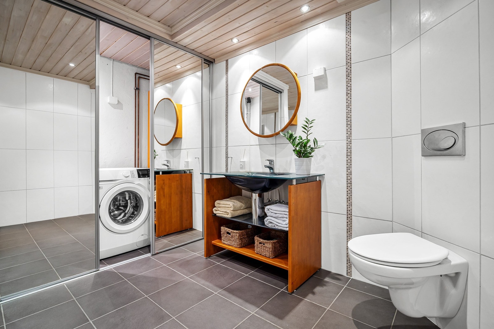 Badet er utstyrt med gulvstående innredning med heldekkende servant, badekar og vegghengt toalett. Bak skyvedør med speilfronter er det veggmontert utslagsvask, varmtvannsbereder og opplegg for vaskemaskin. Galleribilde