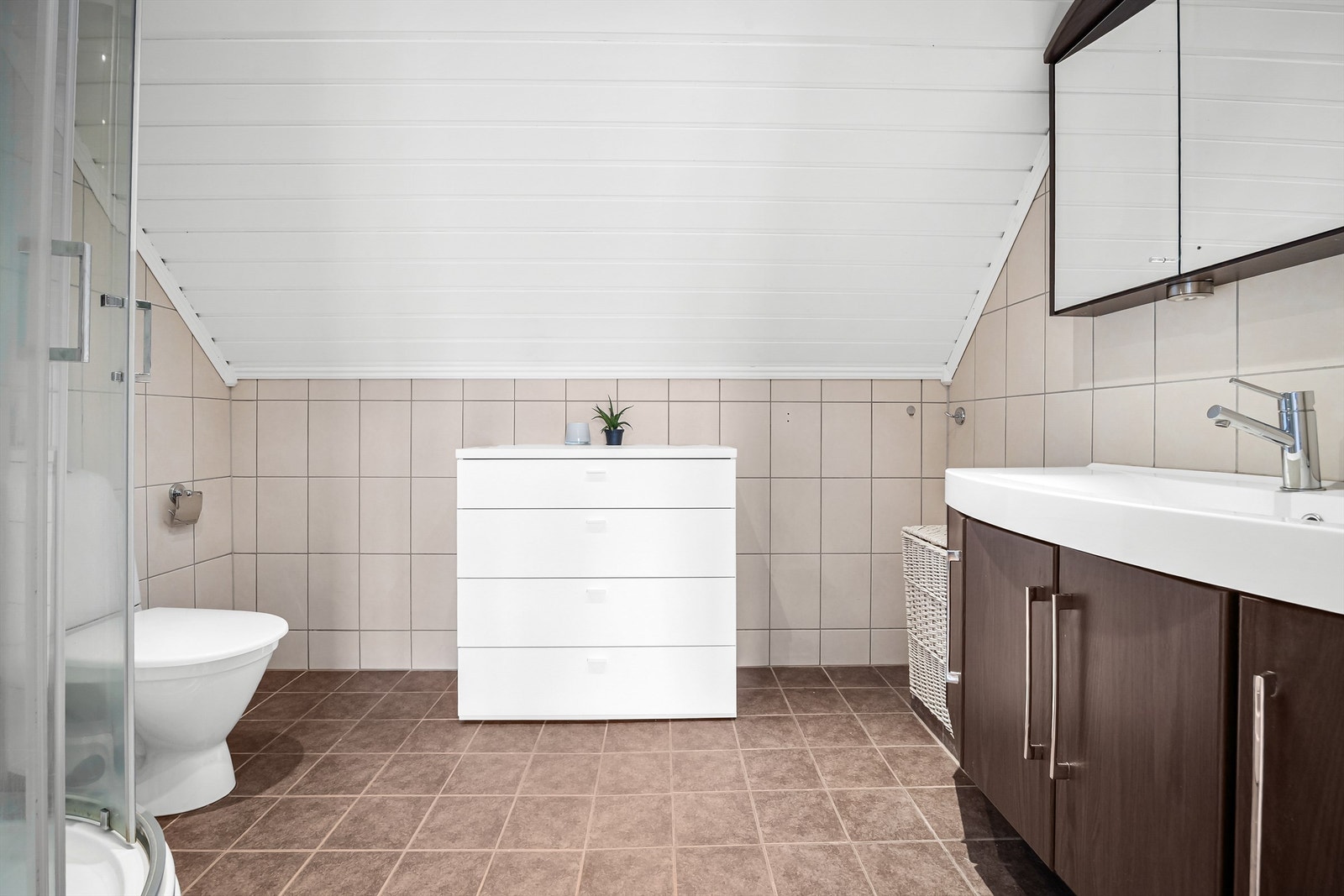 Badet er innredet med vegghengt servantinnredning og speilskap med overlys og stikkontakt på vegg over servant. Videre er badet utstyrt med dusjkabinett med skyvedører og gulvstående toalett. Galleribilde