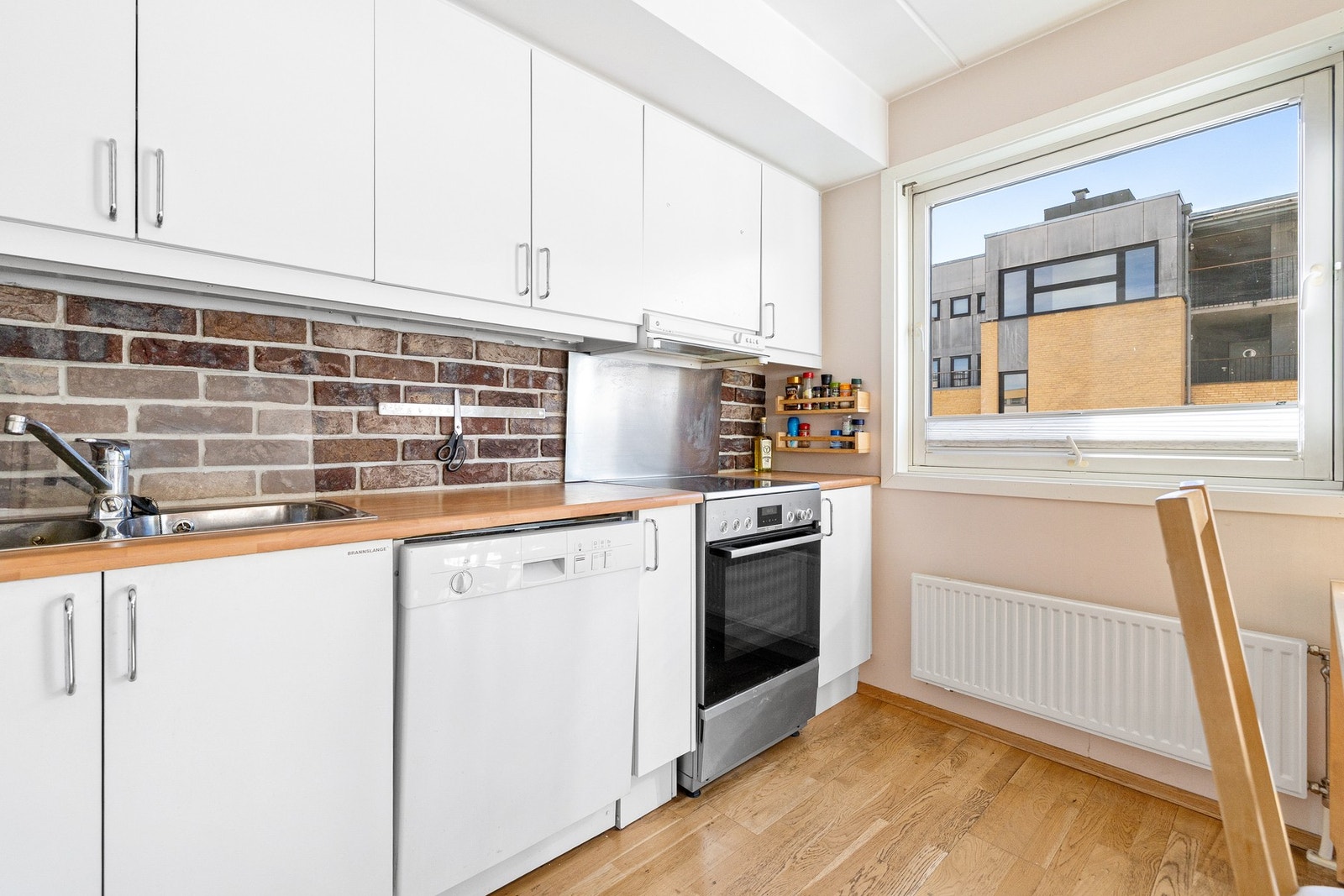 Praktisk kjøkkenløsning med nisjer for komfyr, oppvaskmaskin og kjøl-/fryseskap. Børstet metallplate over kokesonen gir et stilrent uttrykk. Galleribilde