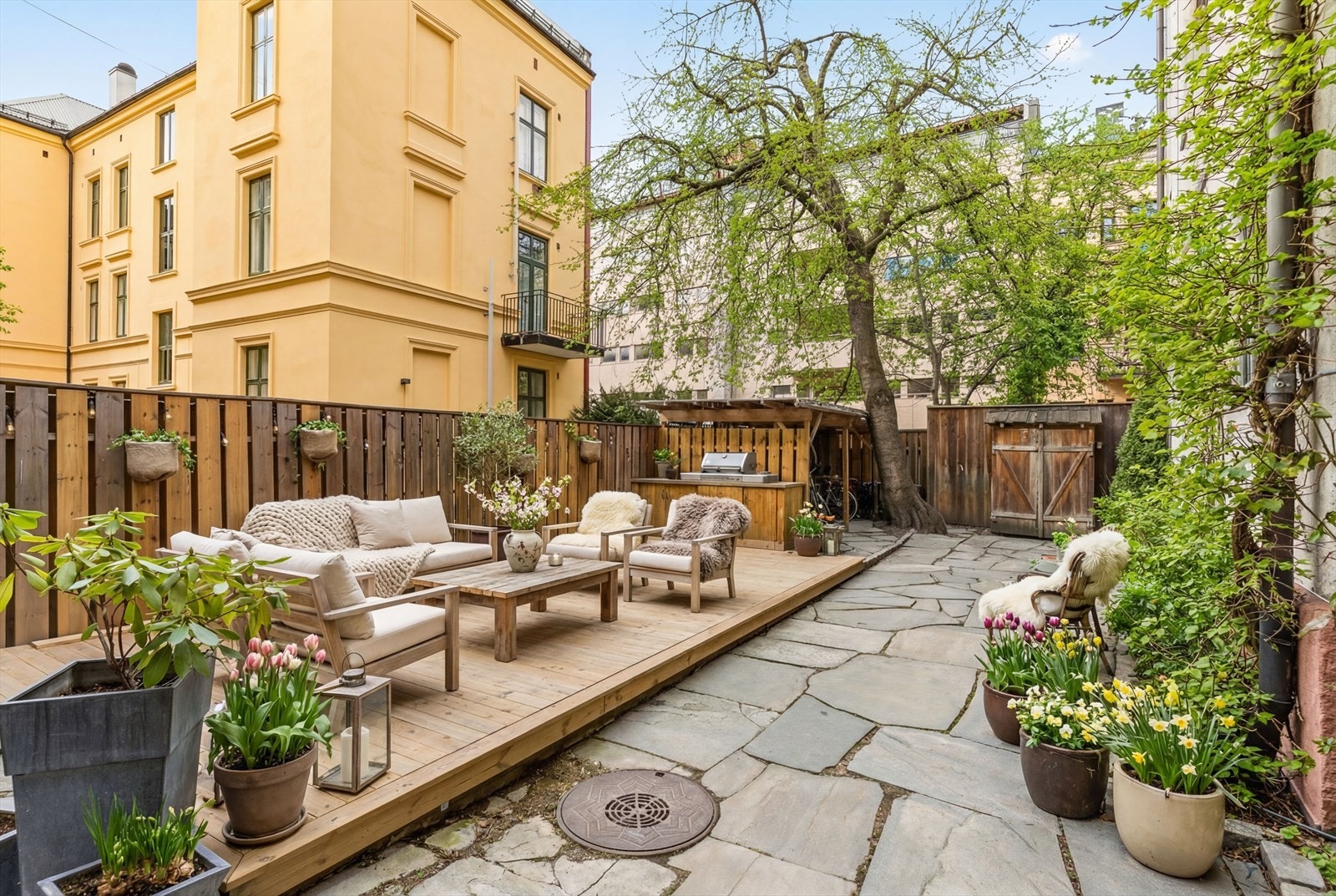 Pent opparbeidet bakgård med steinheller, beplantning og terrasseplatting, utstyrt med sykkelskur og hyggelige utemøbler som kan benyttes av beboerne. Et trivelig uteområde midt i byen. Husdyr er også tillatt. Bilde er til infi AI redigert. Galleribilde