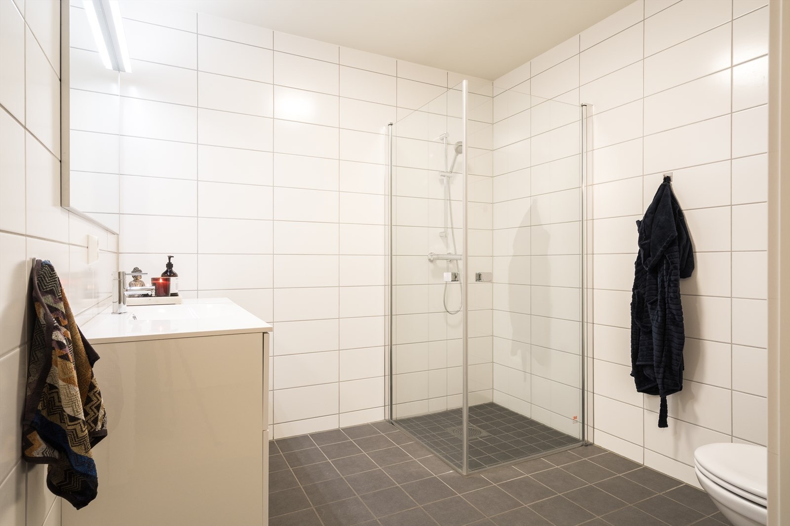 Det er to bad i boligen. Badet nede har en servant med hvite skuffer og et speil med belysning. Galleribilde