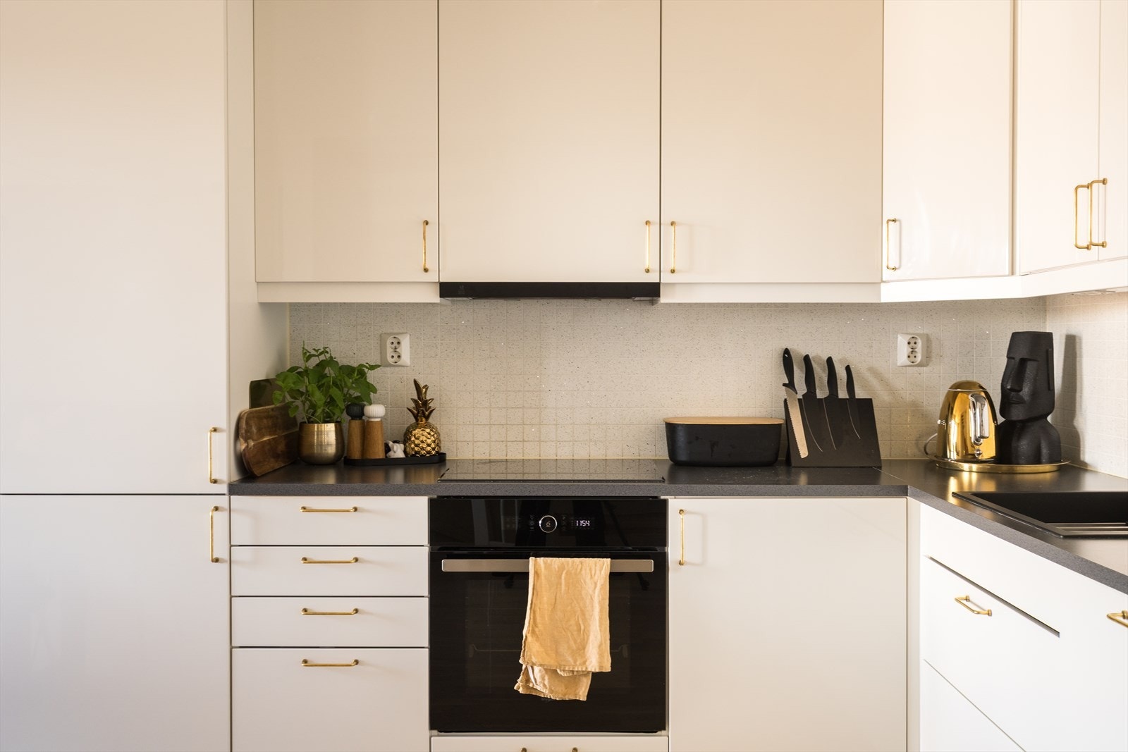 Innredningen har hvite, glatte fronter, gullfargede håndtak, mørk benkeplate av laminat og nedfelt oppvaskkum i sort design. Galleribilde