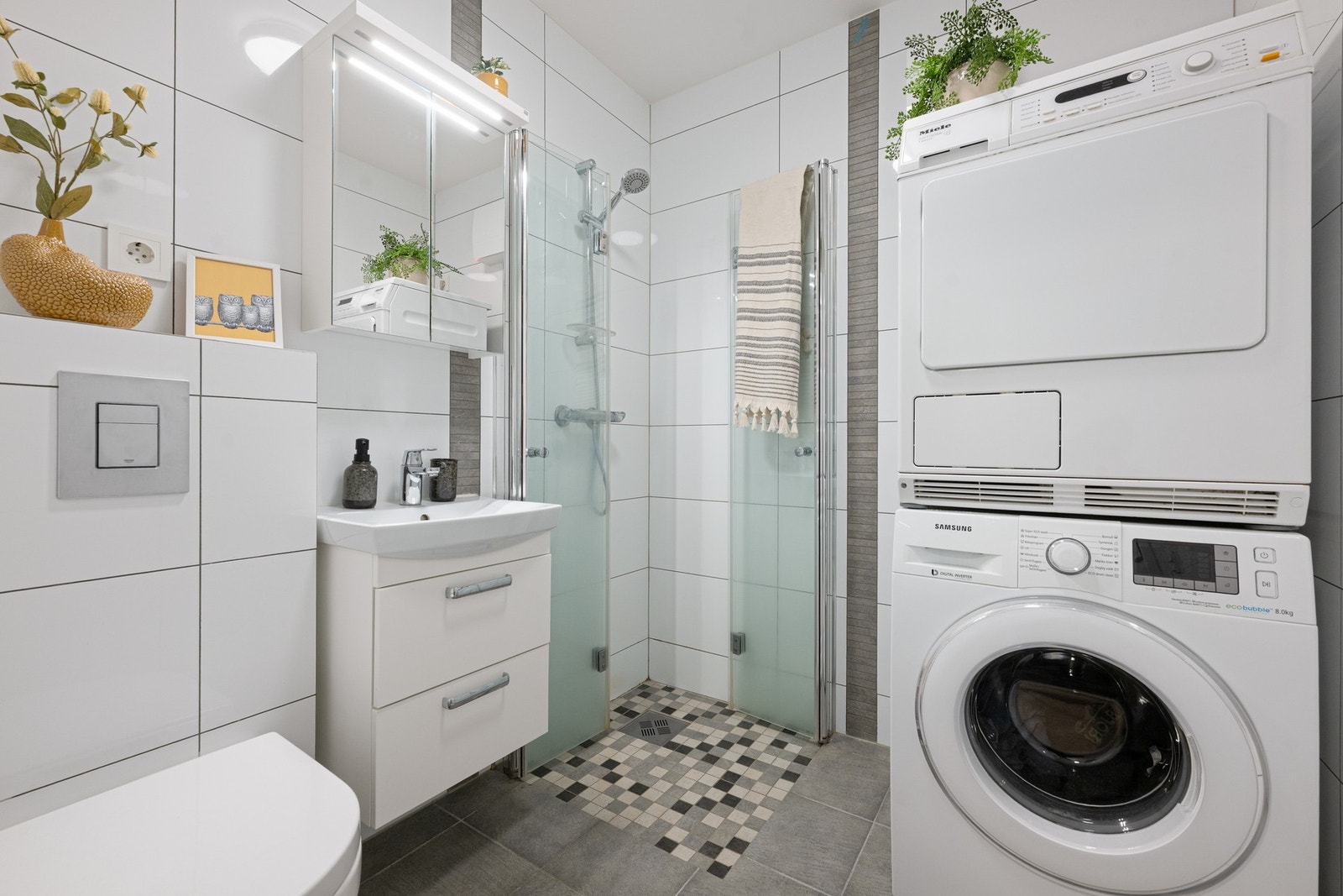 Badet er flislagt og har en praktisk og moderne utforming med servantskap med heldekkende servant, speilskap med lysarmatur, veggmontert toalett og dusjhjørne med foldedører. Galleribilde