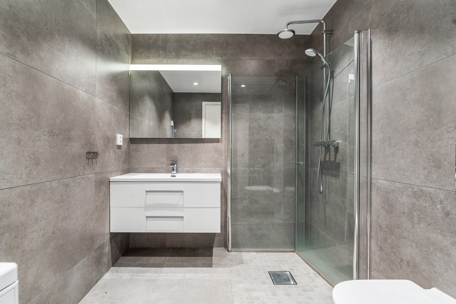 Badet er utstyrt med dusjhjørne med glassvegger, vegghengt toalett og servantinnredning. Galleribilde