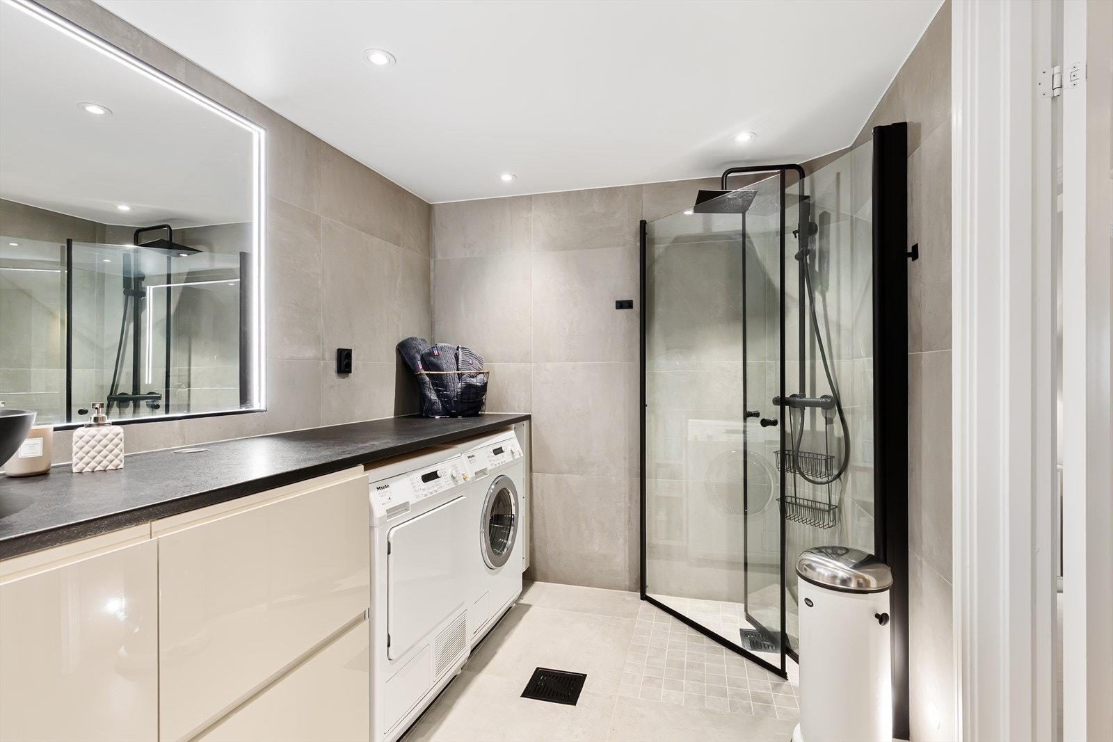 Badet fremstår delikat og moderne, helfliset med store beige fliser som gir et tidløst og eksklusivt uttrykk. Dusjsonen er utstyrt med stilrene glassvegger og sorte detaljer som gir et moderne preg. Galleribilde
