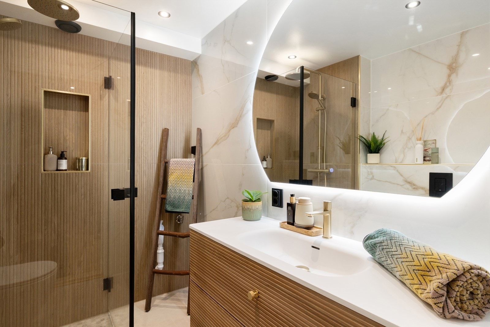 Badet fremstår stilrent med eksklusive materialvalg, hvor kombinasjonen av lyse fliser og varme tretoner gir et elegant uttrykk. Innredningen består av servant med ettgreps armatur, romslig skuffeløsning og speil med integrert belysning. Galleribilde