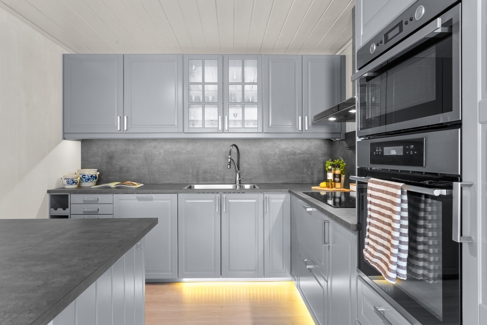 Kjøkkenet består av grå, profilerte fronter samt ett overskap med vitrinedører, laminat benkeplate med underlimt kum og ett-greps blandebatteri. Galleribilde