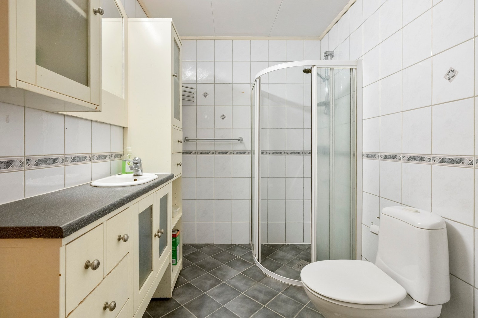 Flislagt bad innredet med vegghengt baderomsmøbel med vask, frittstående toalett og et dusjhjørne med dusjkant og glassvegger. Galleribilde