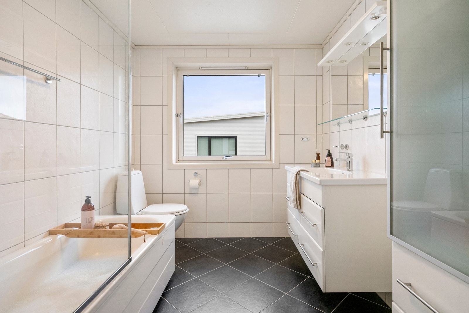 Bad innredet med heldekkende servant montert på en vegghengt skuffeseksjon, speil med belysning og frittstående toalett. I tillegg er rommet utstyrt med badekar. Galleribilde