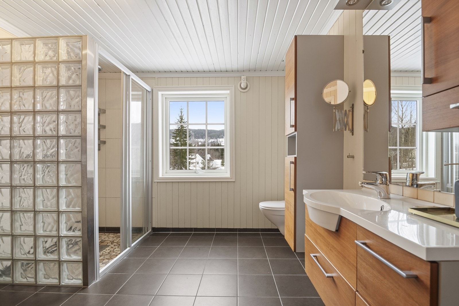 Badet er utstyrt med dusjhjørne, vegghengt toalett, veggskap, speilskap, servant med underskap og opplegg til vaskemaskin. Galleribilde