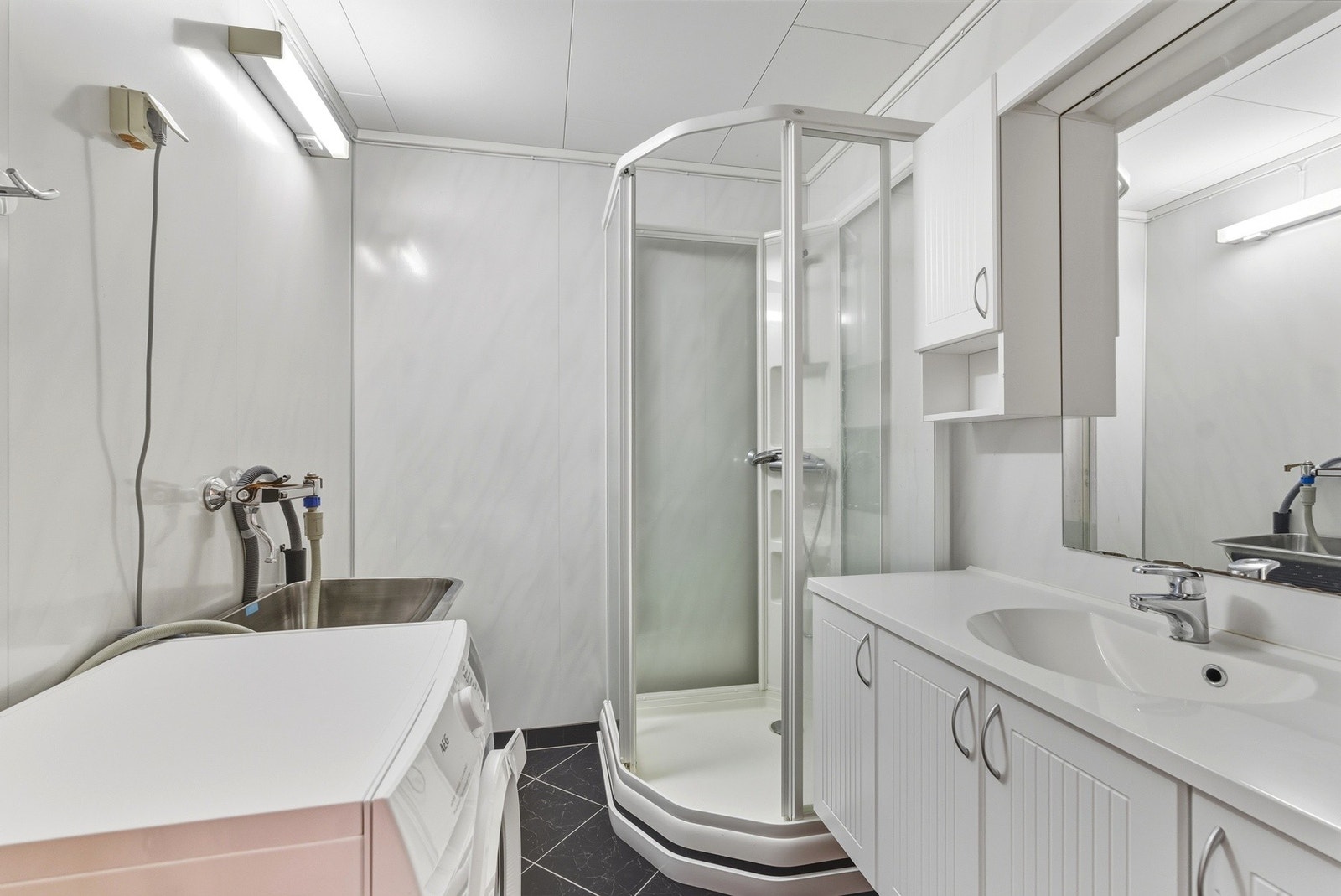 Badet har dusjkabinett, veggmontert toalett, opplegg for vaskemaskin, skyllekar i stål montert på vegg og helstøpt servantinnredning med lys over speilet. Galleribilde