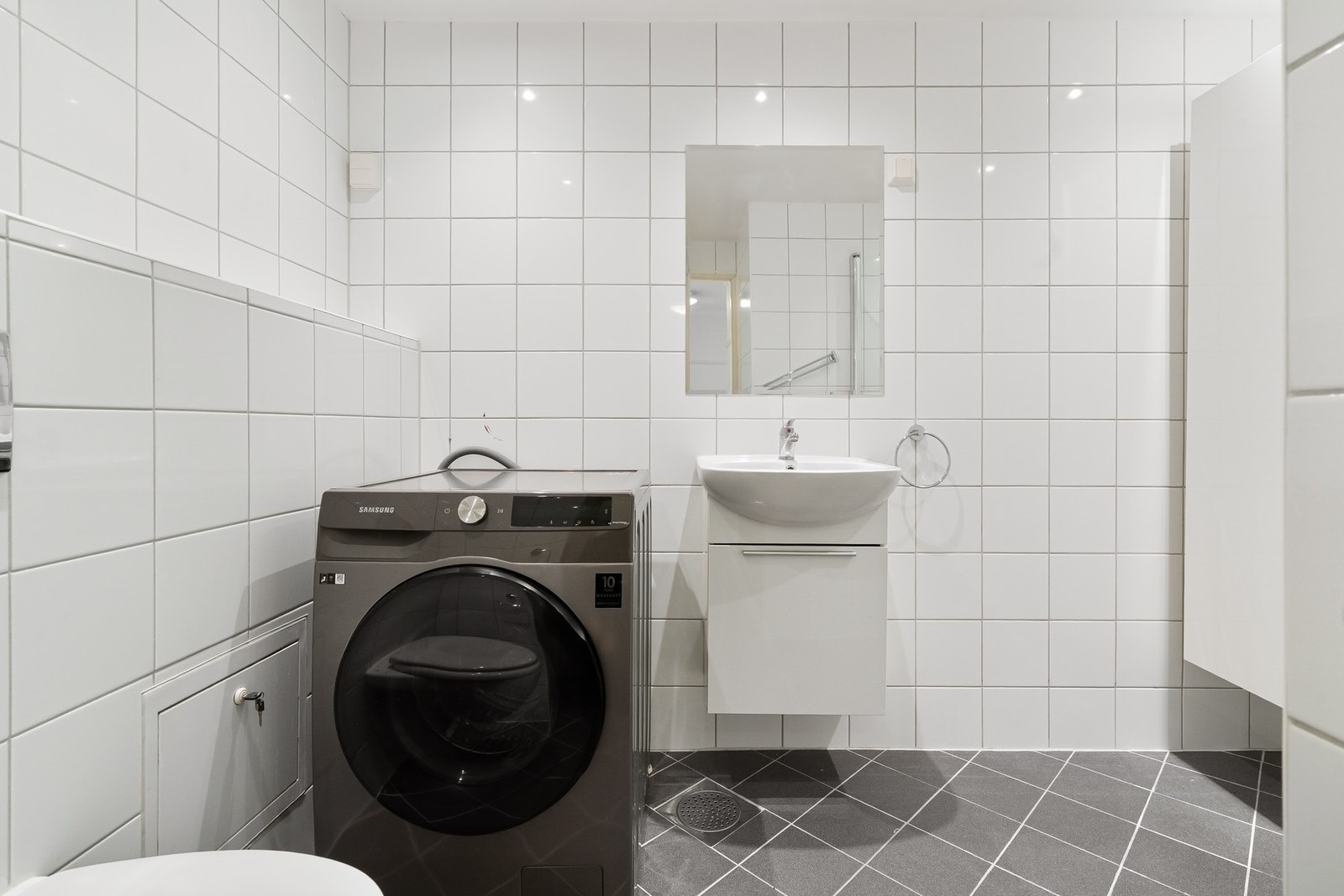 Badet er flislagt med servant i baderomsinnredning, høyskap, dusjløsning med frittstående glassvegger, vegghengt toalett og tilkobling for vaskemaskin. Galleribilde