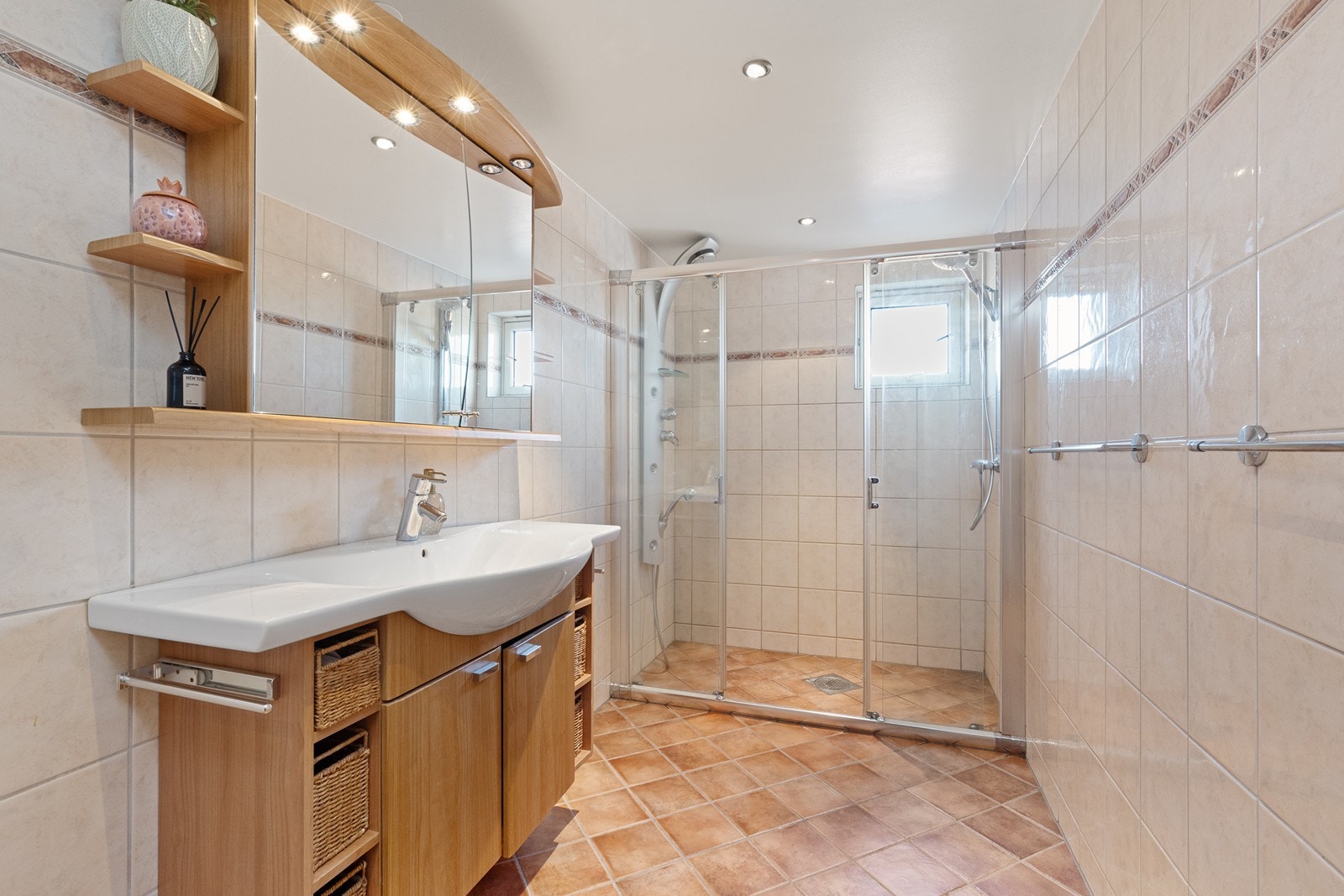 Badet er utstyrt med veggmontert toalett, dusjnisje med skyvedører. Innredningen er trefarget og har glatte fronter, flere åpne hyller, heldekkende servant og speilskap med belysning i overkant. Galleribilde