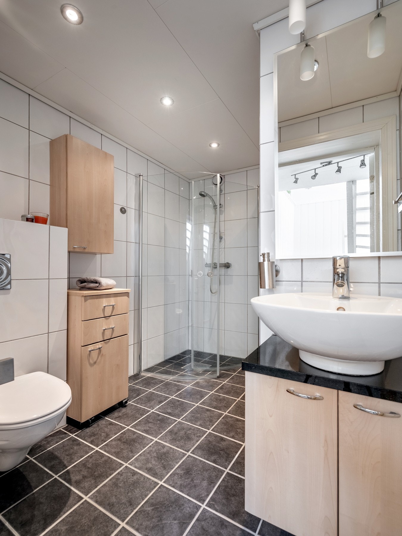 Badet i underetasjen har flislagte vegger og flislagt gulv med elektriske varmekabler. Innredningen består av servant på skapseksjon, speil over servant, veggmontert toalett og dusjvegger/hjørne. Galleribilde
