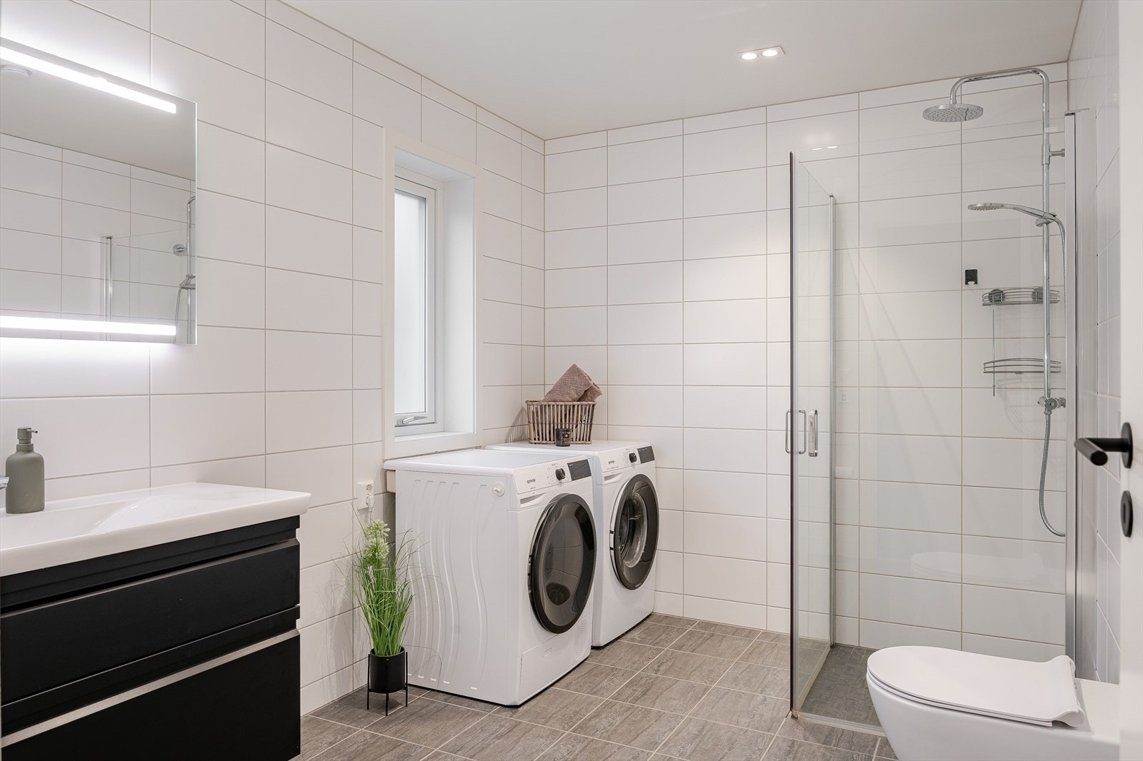 Baderommet er utstyrt med dusjhjørne med innfellbare glassdører og både hånd- og regnfallsdusj, samt vegghengt toalett. Det er også god plass til både vaskemaskin og tørketrommel. Galleribilde