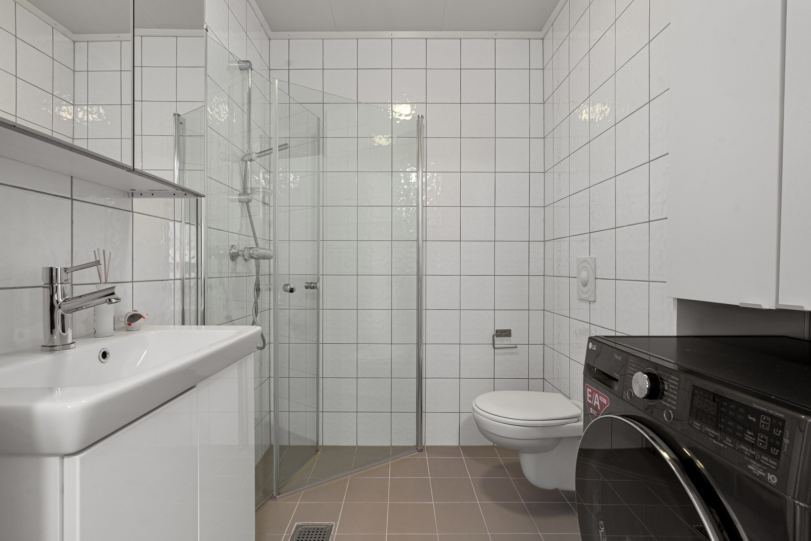 Innbydende flislagt baderom innredet med servant, dusjvegger av glass og dusjgarnityr på vegg, samt opplegg for vaskemaskin