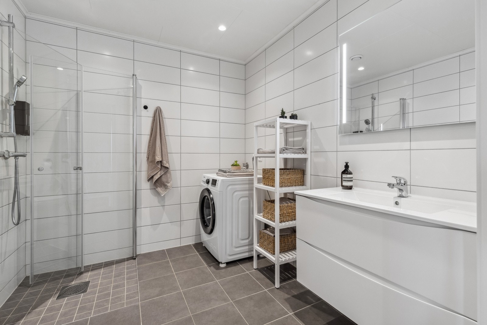 Videre har badet en nedsenket dusjsone med mindre fliser på gulv, innfellbare glassdører og hånddusj. Det er avsatt plass samt opplegg for vaskemaskin og tørketrommel. Himlingen er pent lyssatt med LED downlights. Galleribilde