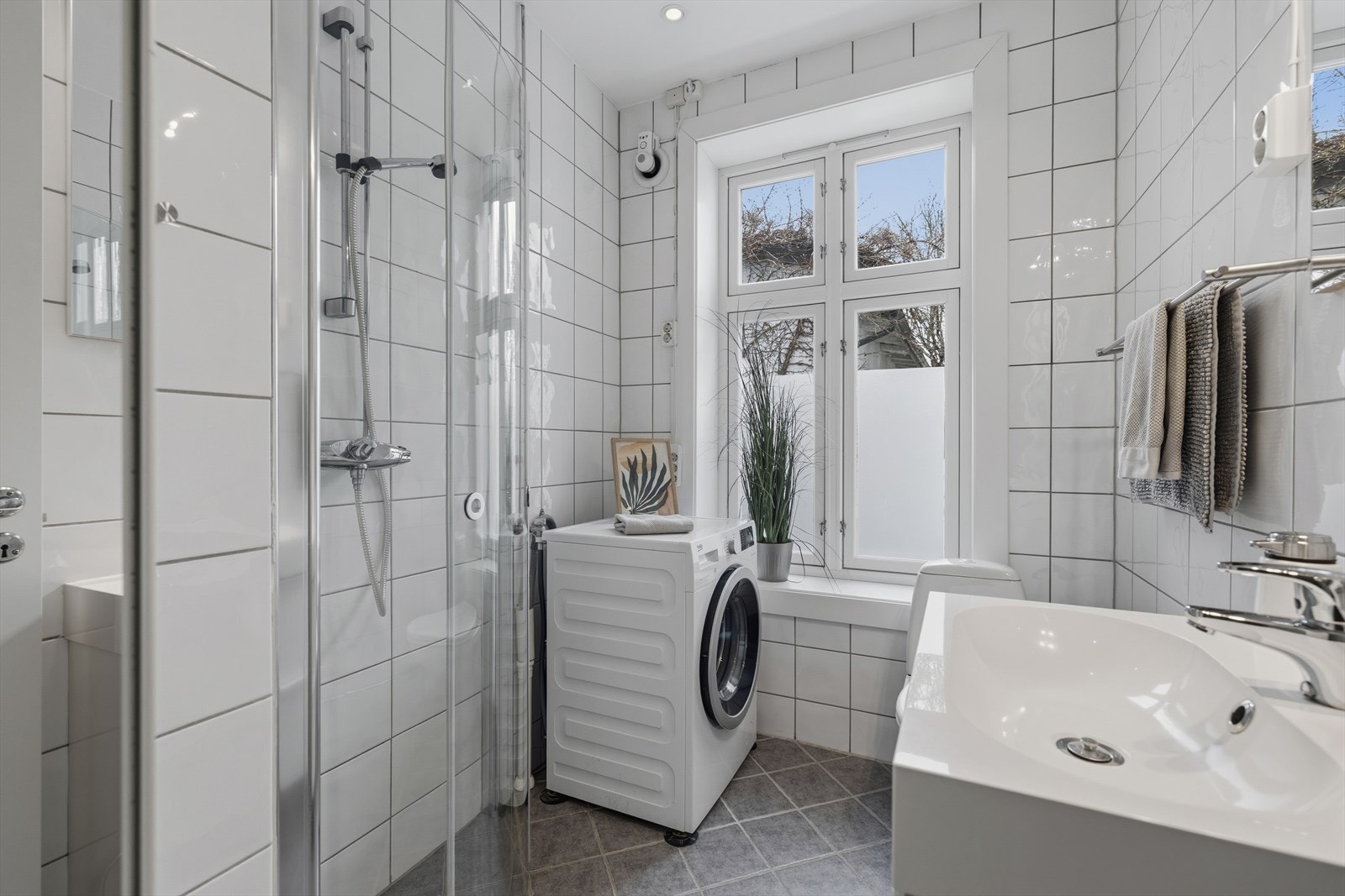 Lyst og tidløst bad med fliser på vegger og gulv. Det er opplegg for vaskemaskin i hjørnet. Galleribilde