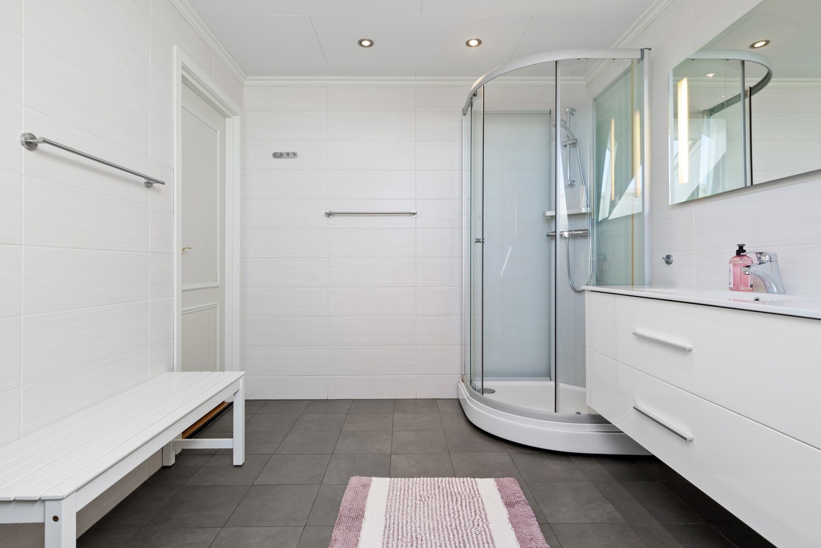 Badet er utstyrt med dusjkabinett, vegghengt toalett og servant med tilhørende innredning. Det er varmekabler i gulvet og downlights i taket. Galleribilde