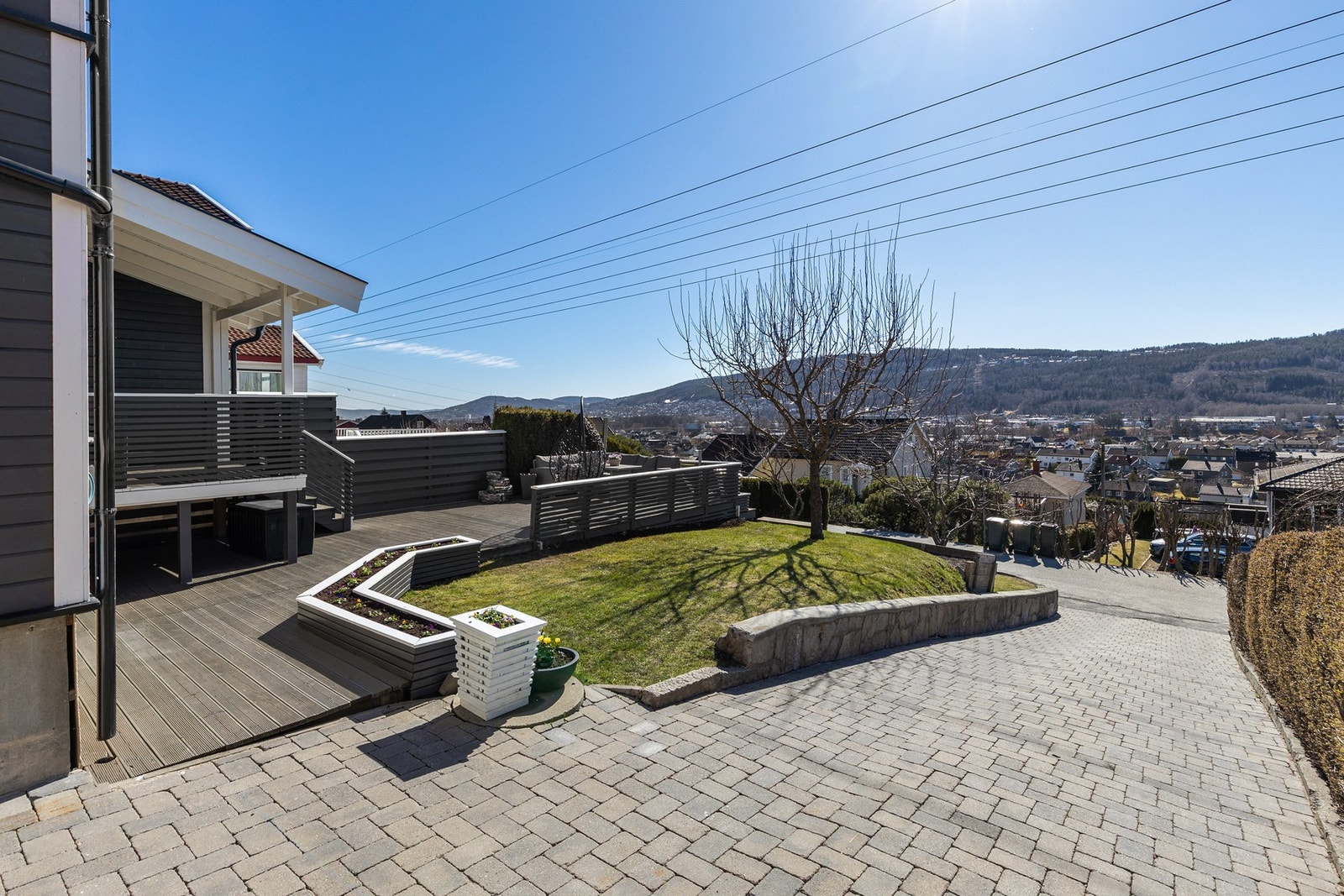 Steinbelagt gårdsplass og pent opparbeidet tomt med gressplen, trær og platting/terrasse. Galleribilde