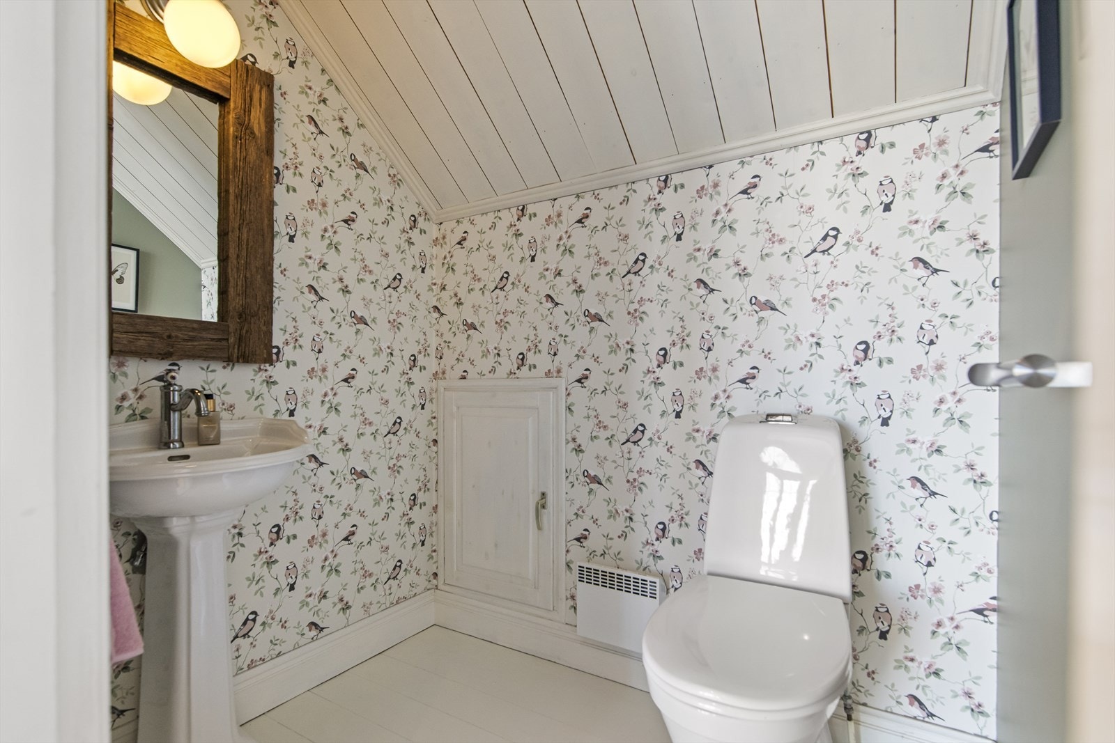 Wc-rommet har malt tregulv, en elegant søyleservant og et speil med tilhørende lys. På veggene er det en elegant tapet med fugle- og blomstermotiv. Galleribilde